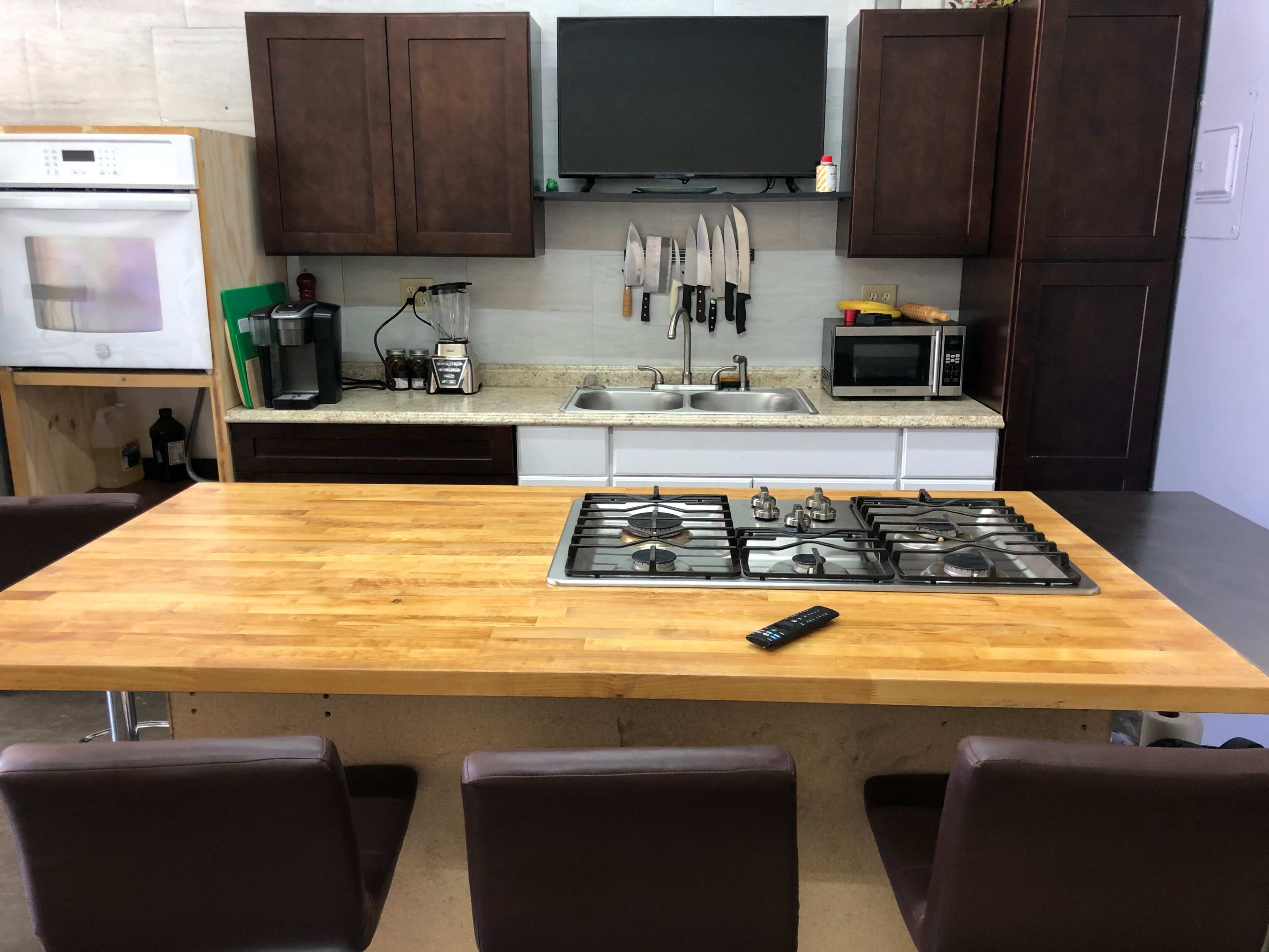 The image shows a kitchen with dark cabinetry, a countertop with a built-in gas stove, and a central wooden table surrounded by four brown chairs.