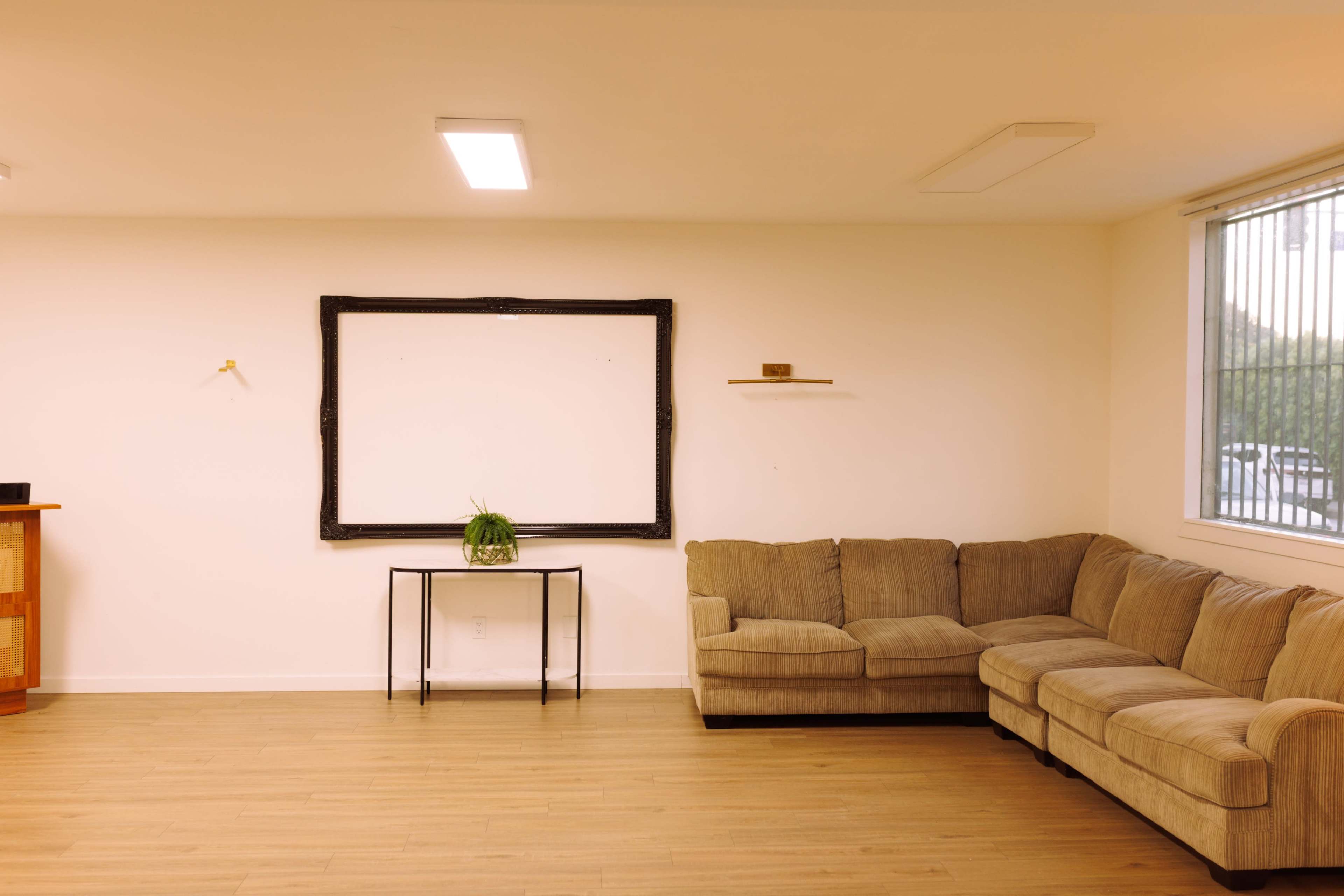 The image shows a minimalist interior space with a beige sectional couch, a small table, and a large empty frame mounted on a white wall.