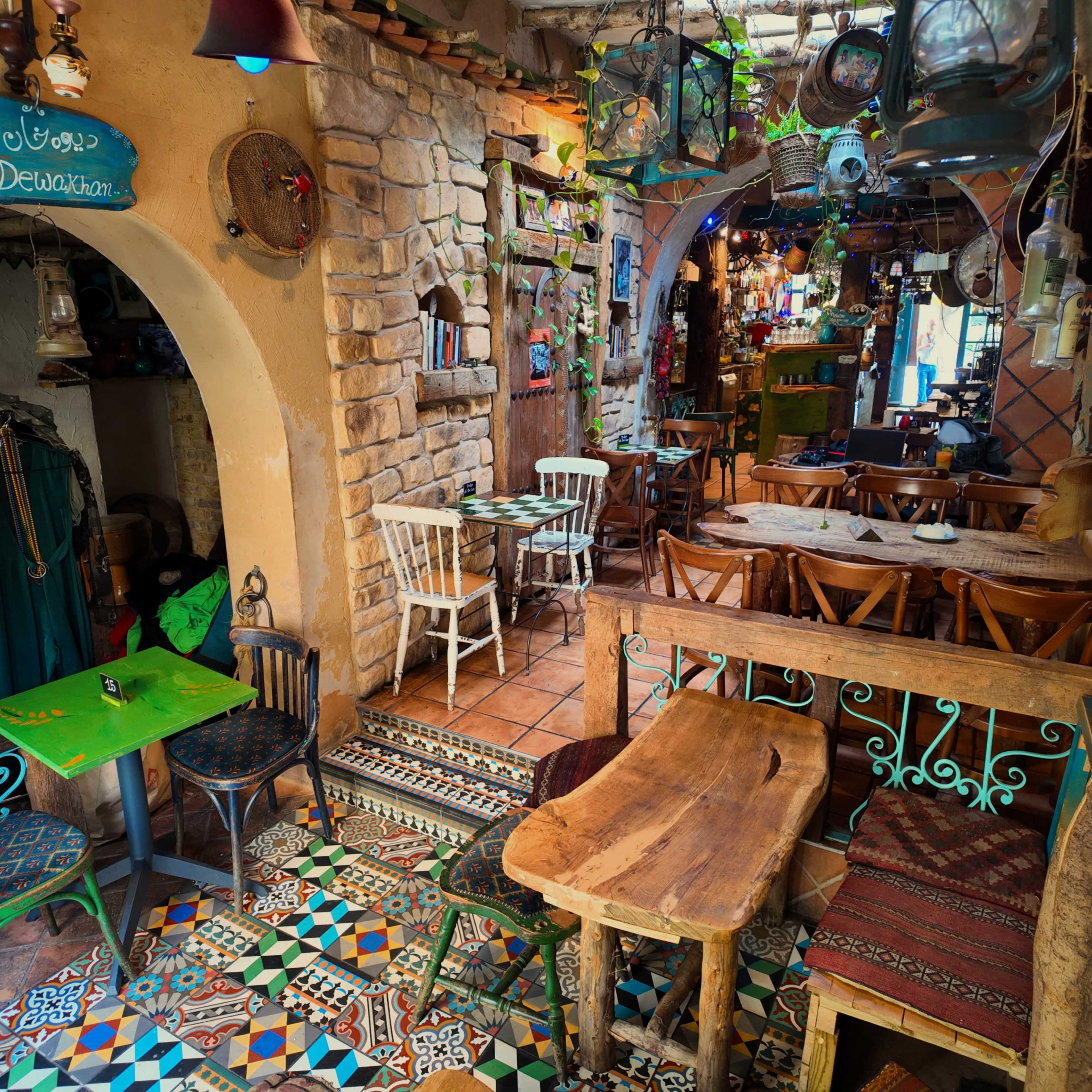 The image shows a colorful, rustic interior of a café or restaurant with wooden tables, ornate tiled floors, and various decorative items hanging from the walls.