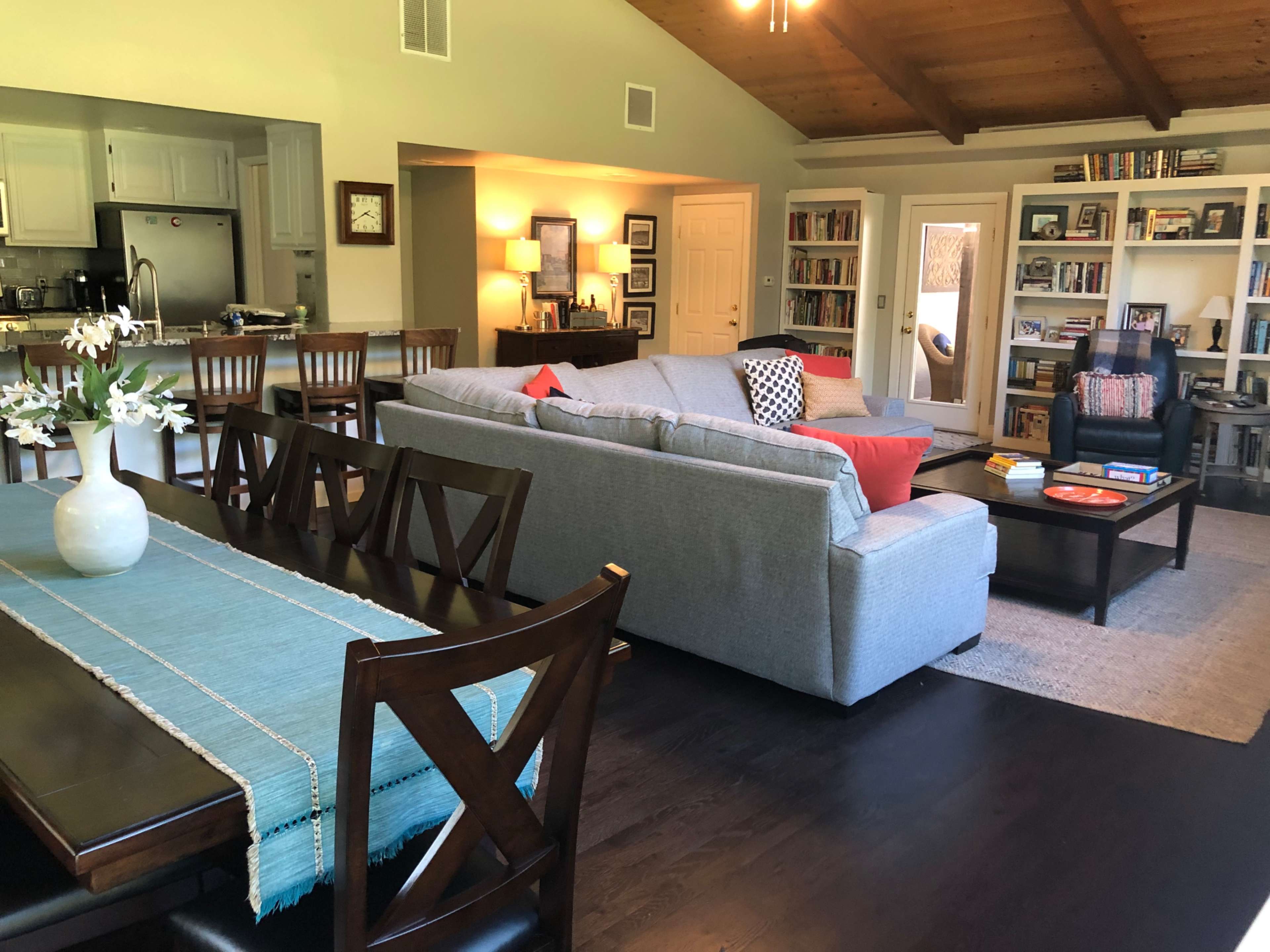 A spacious living room and dining area with a gray sofa, wooden dining table, and shelves filled with books.