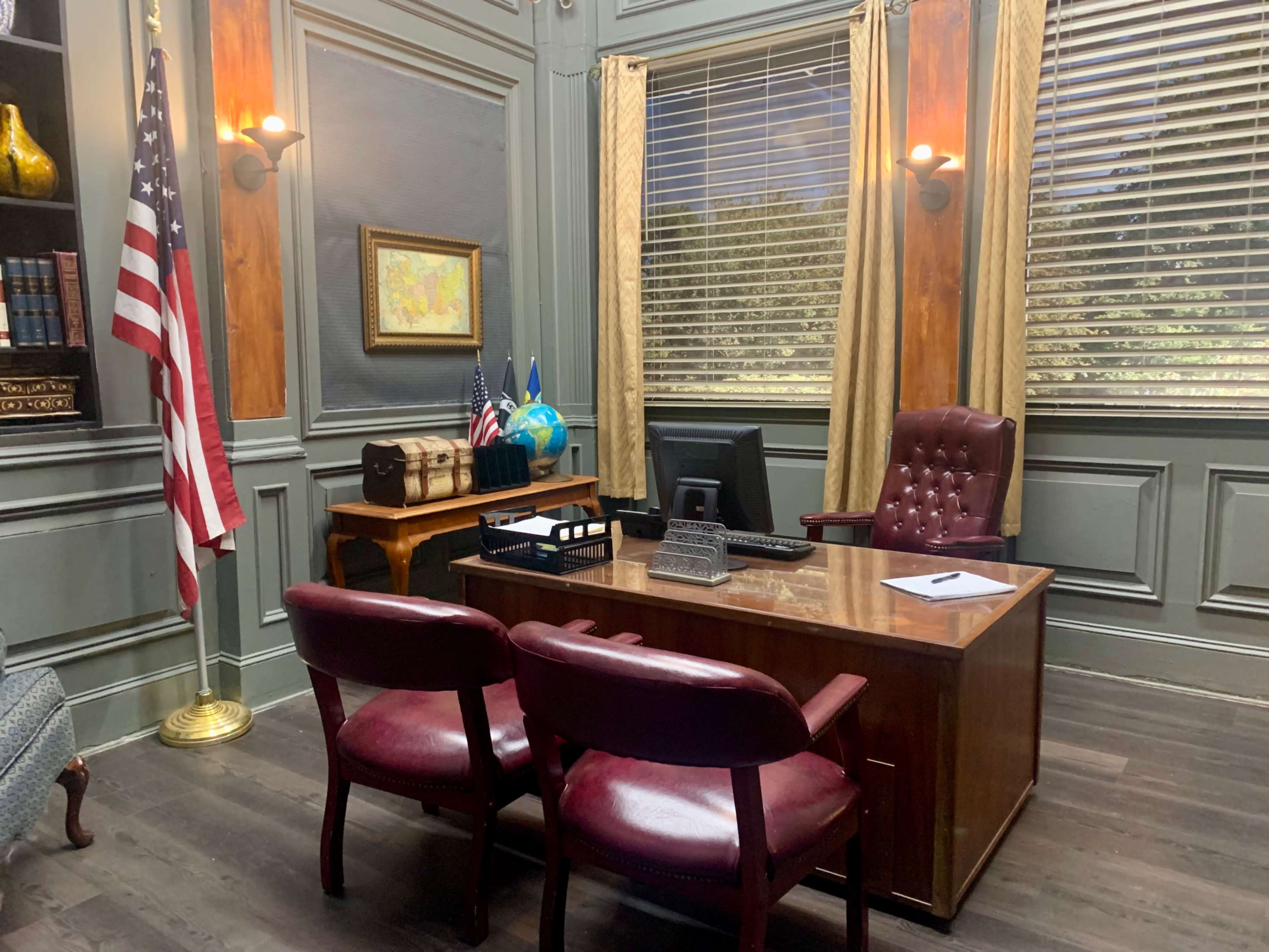 An office with a wooden desk, two maroon chairs, a computer, and flags positioned nearby.