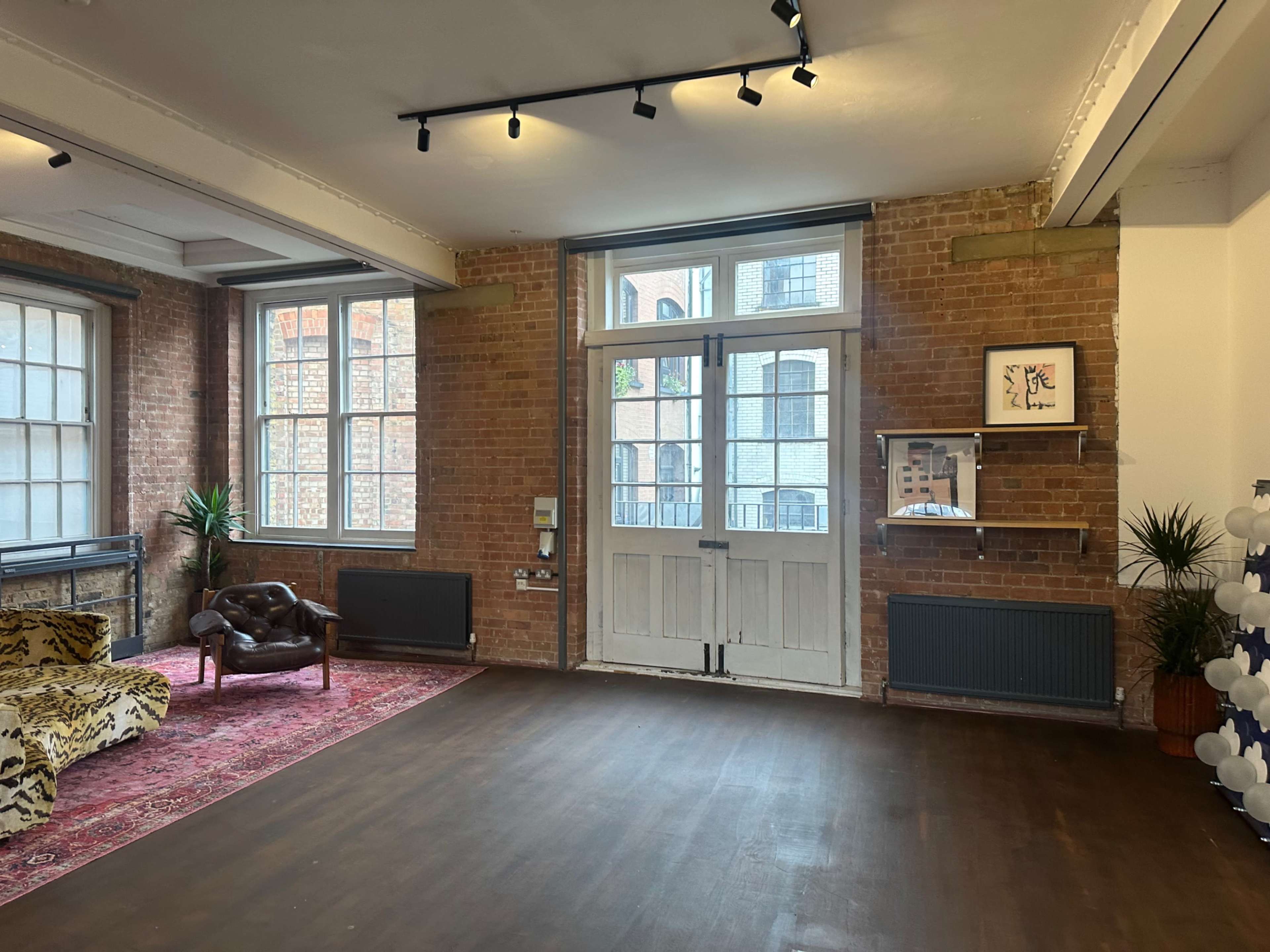 The image shows a spacious room with brick walls, large windows, a patterned sofa, and a wooden floor.