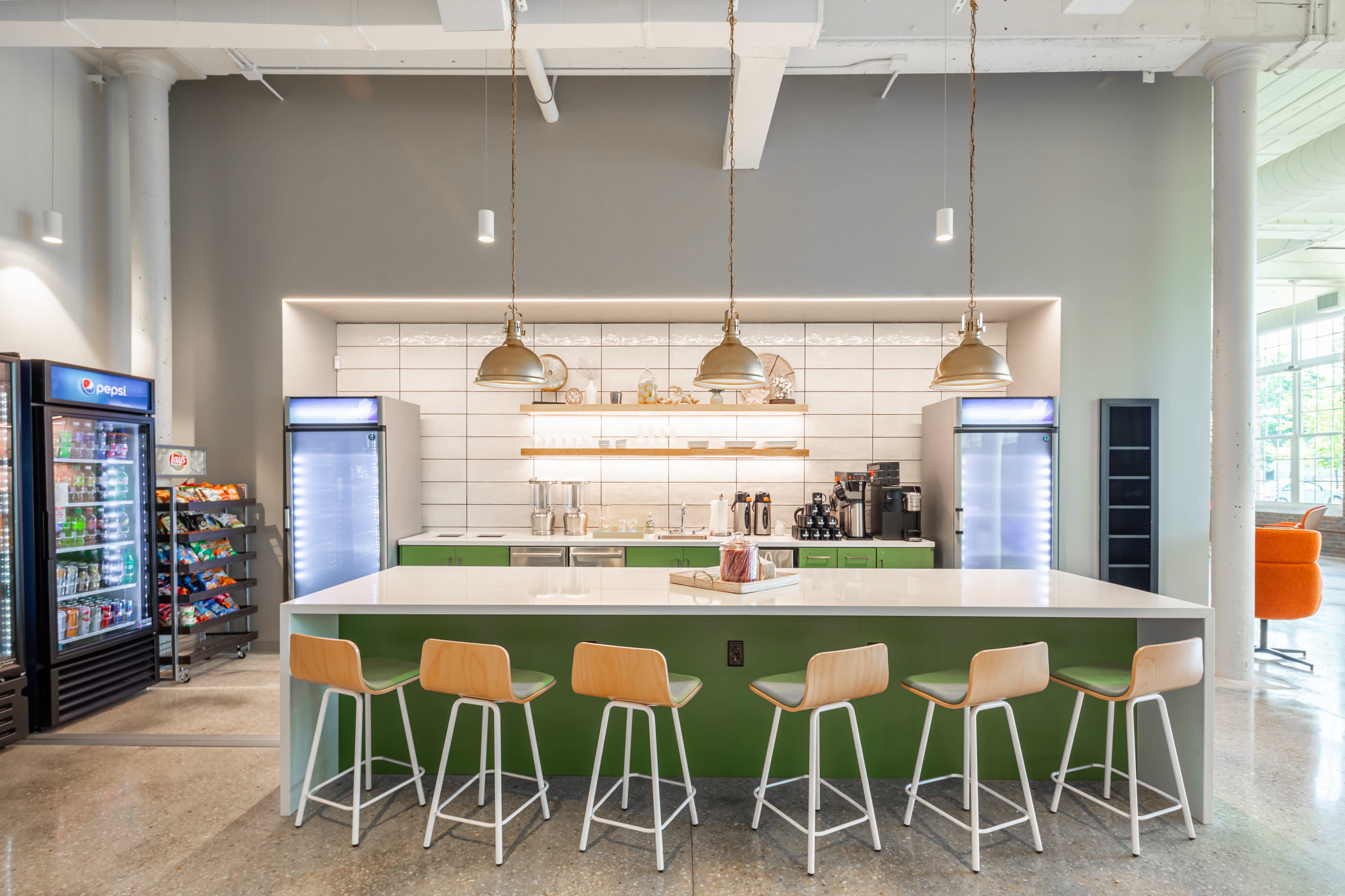 A modern kitchen area with a central island, bar stools, and appliances, including a refrigerator and coffee machines, set against a light-colored wall.