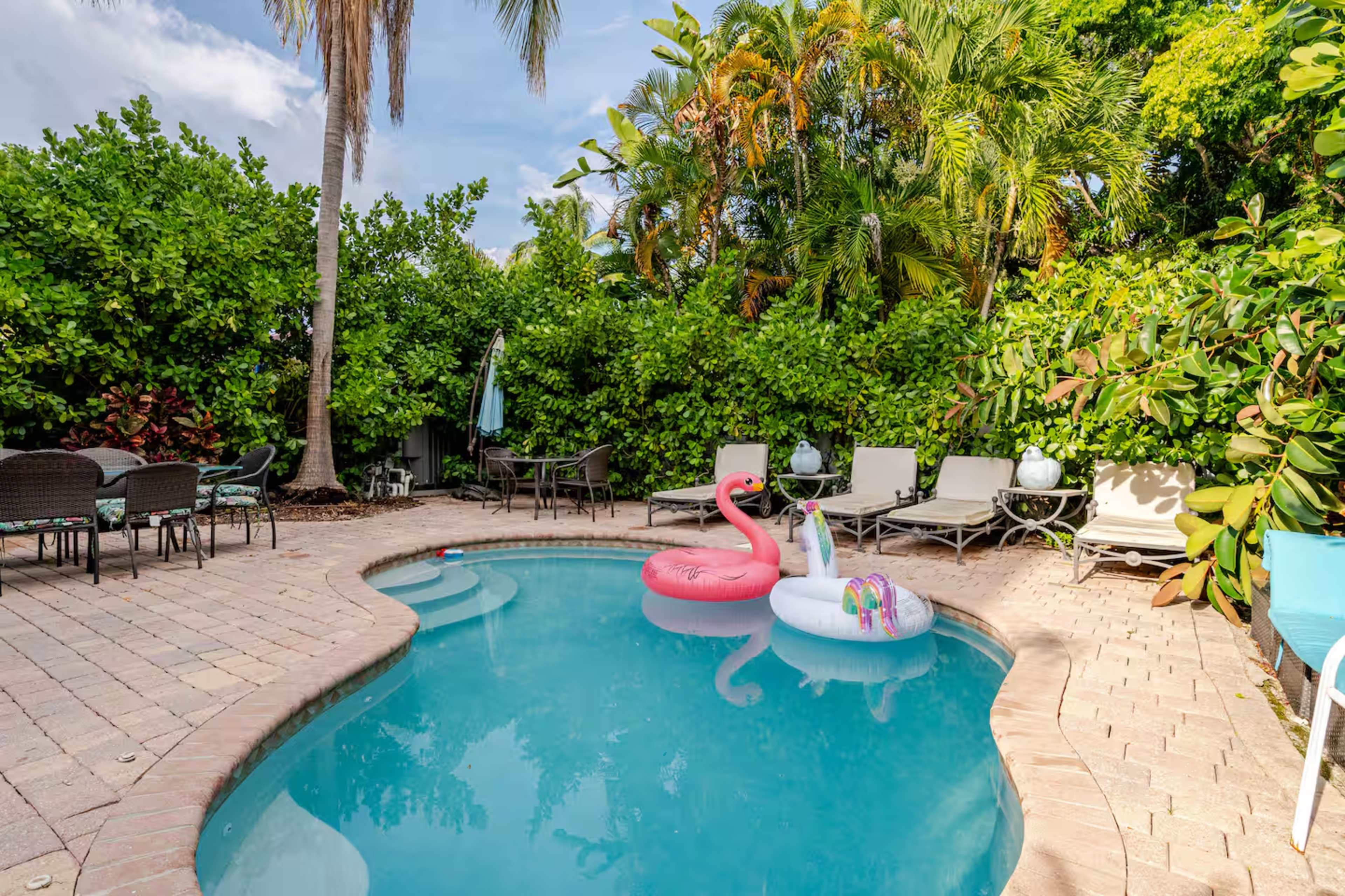 A swimming pool surrounded by lush greenery, with pink flamingo and unicorn floaties and several lounge chairs nearby.