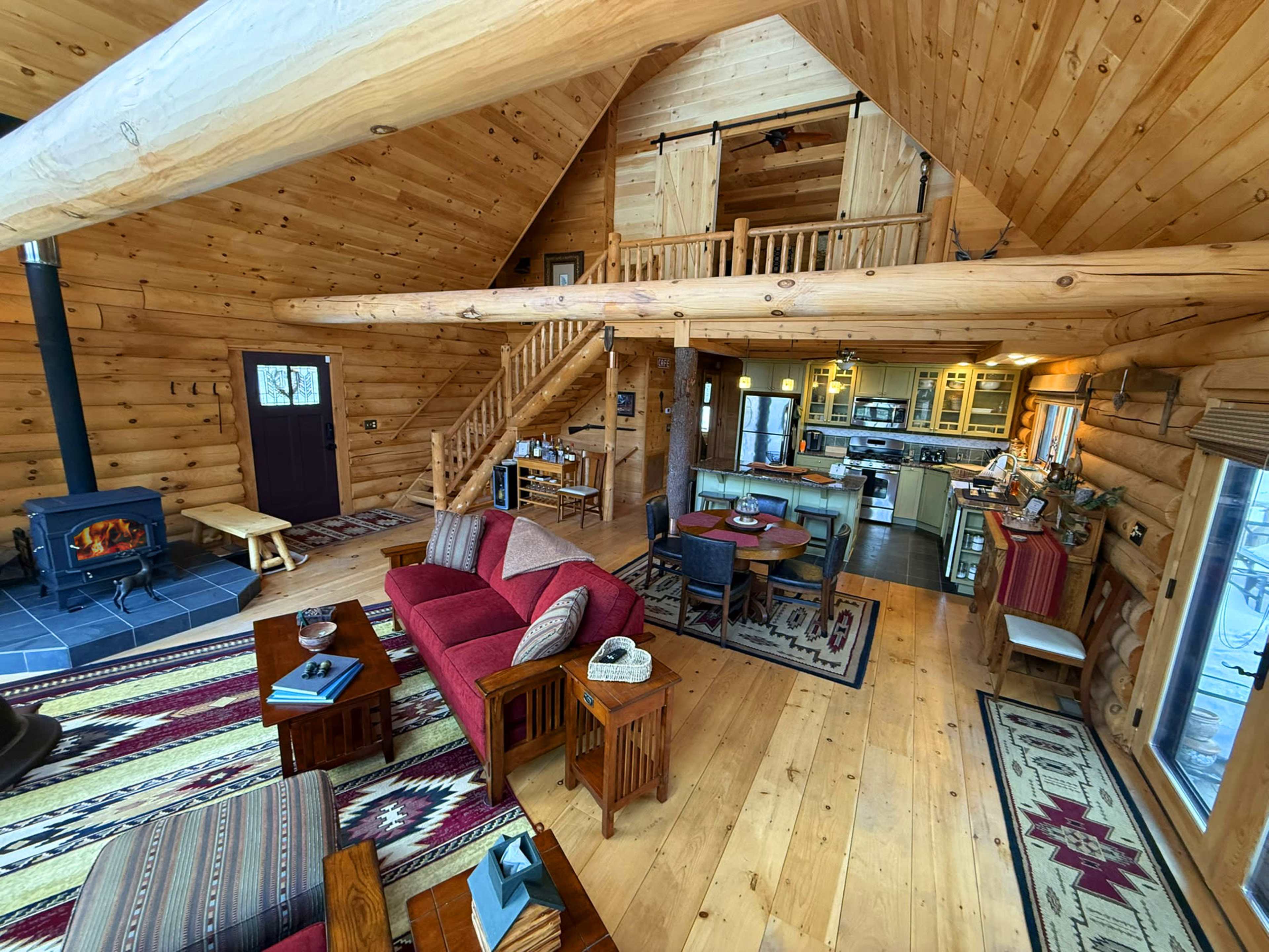 The image shows the interior of a log cabin with wooden walls, a staircase, a fireplace, and a kitchen area in an open layout.