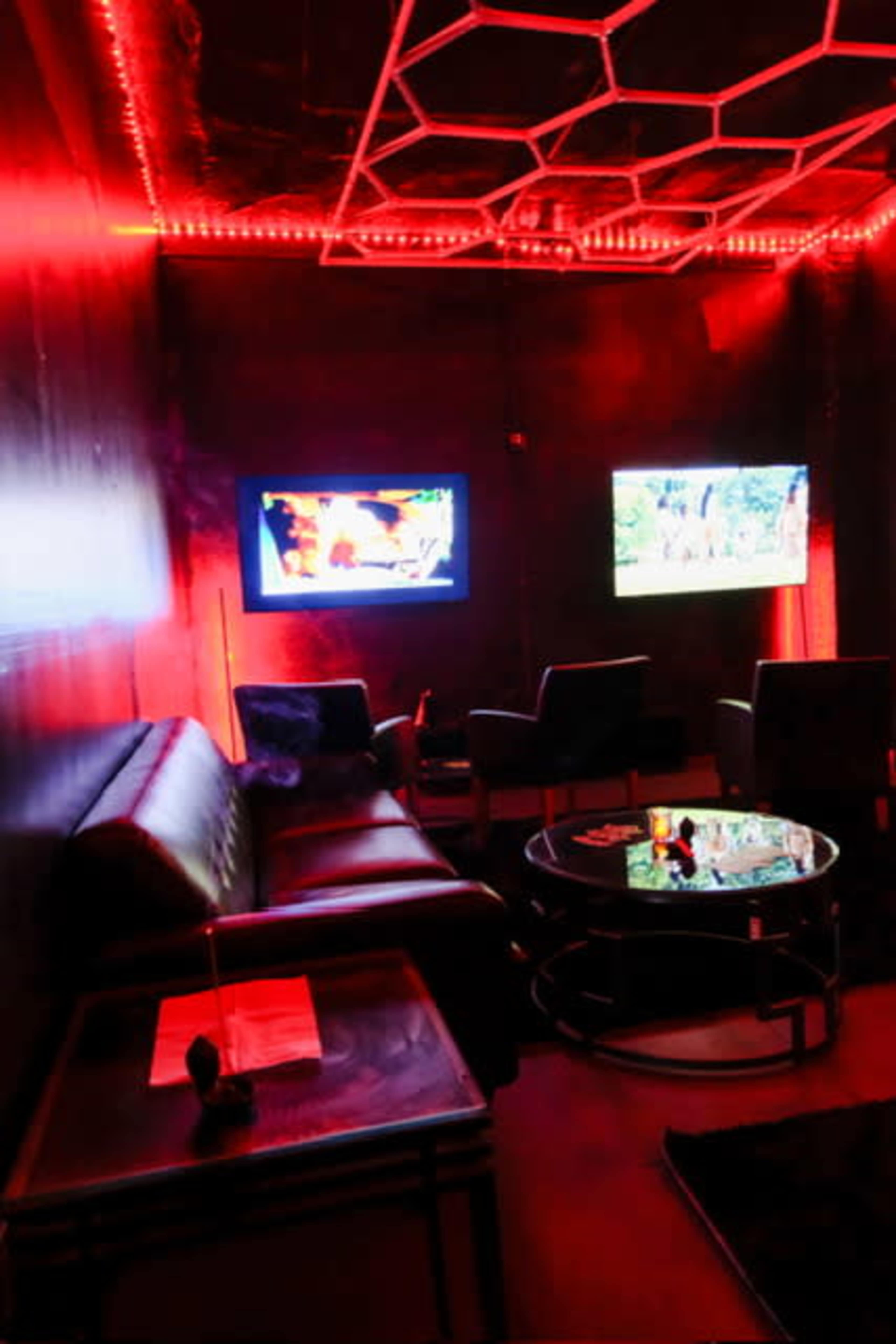 A small, dimly lit lounge area features two televisions, a black leather sofa, and a glass coffee table under red ambient lighting.