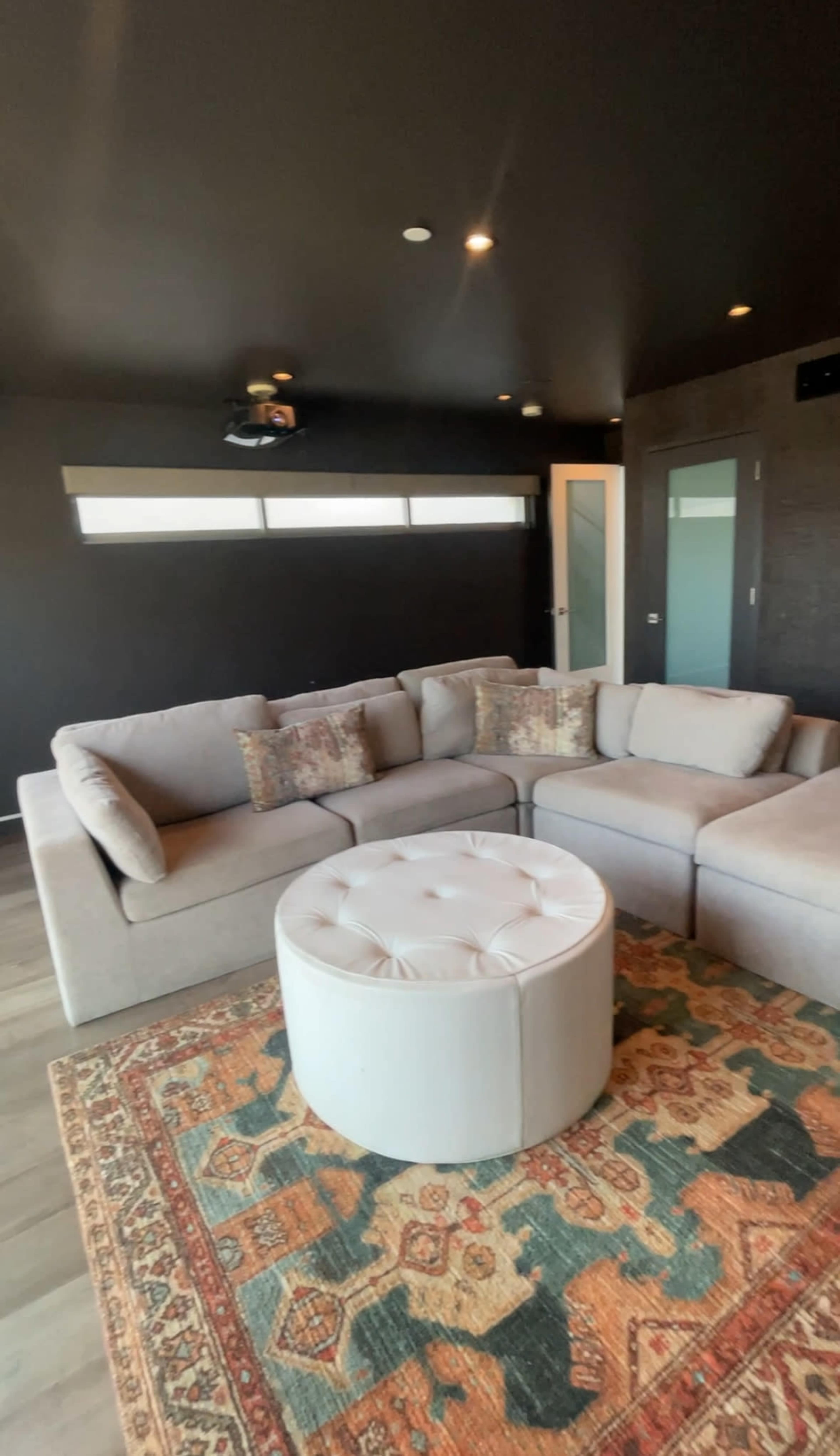 The image shows a modern living room with a grey sectional sofa, a circular white ottoman, and a patterned area rug on the floor.