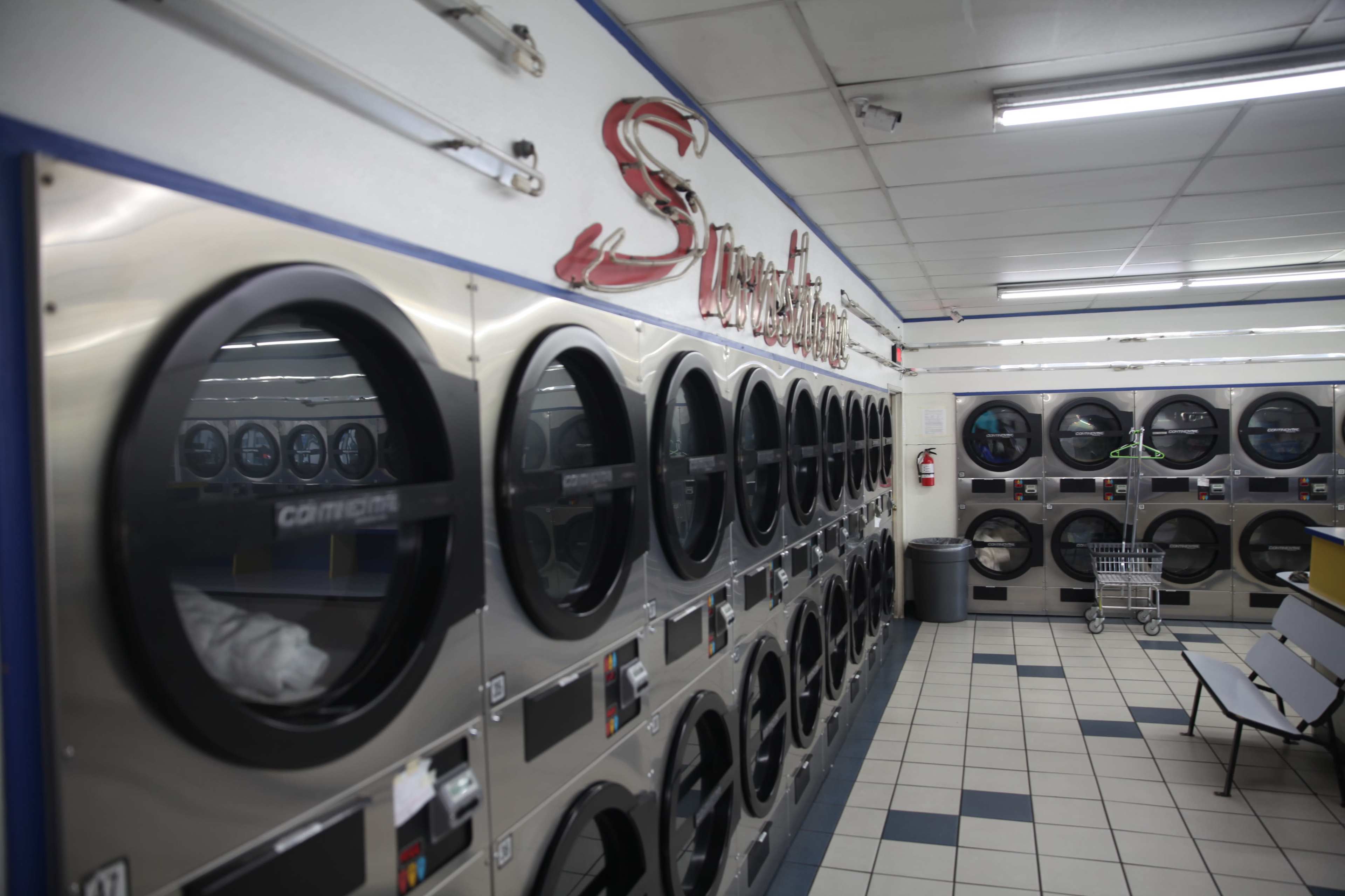 The image shows a laundromat interior with rows of stainless steel washing machines and dryers along the wall.