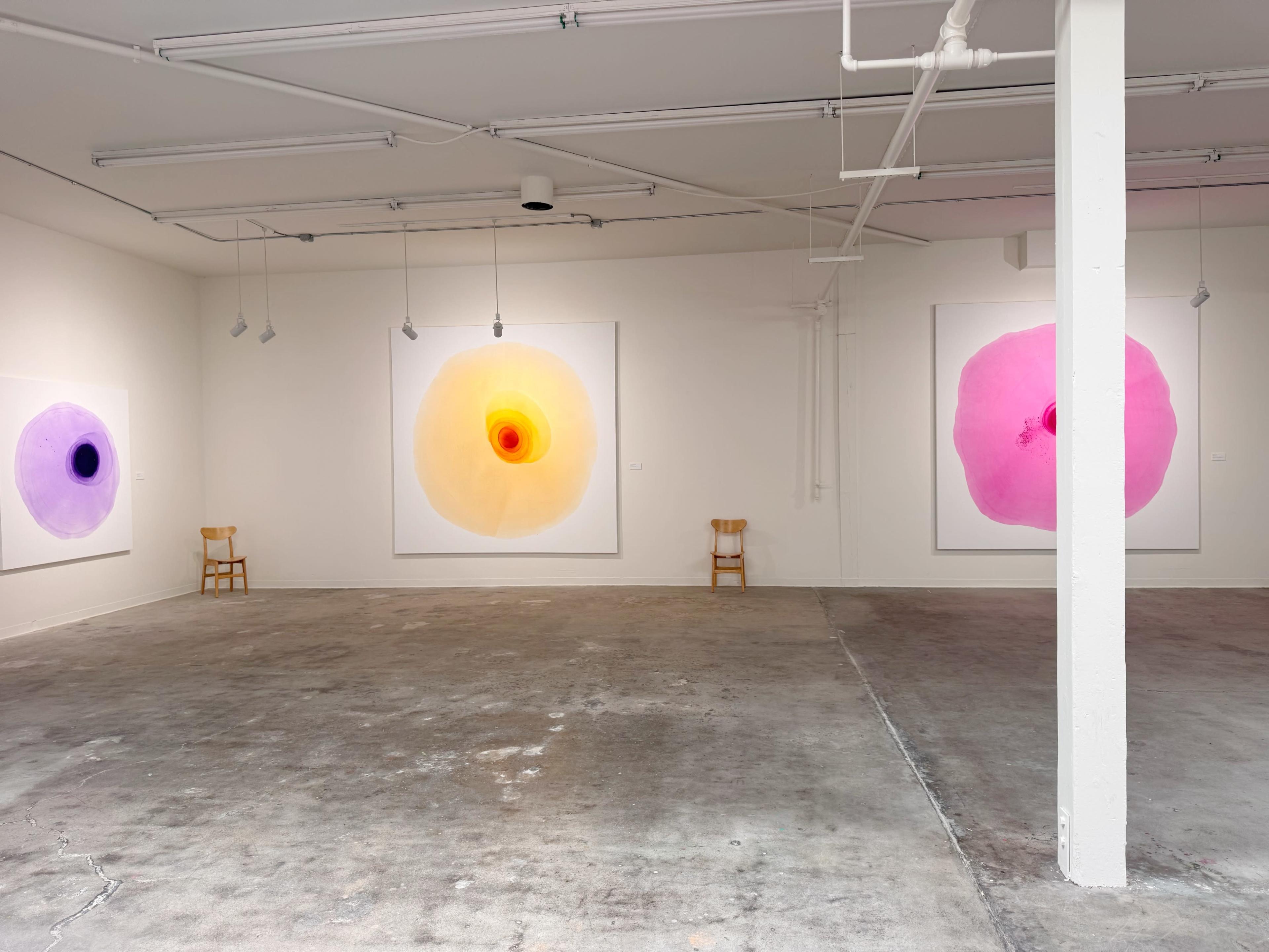 The image shows an art gallery with three large circular paintings in yellow, pink, and purple on the walls, and two wooden chairs positioned on the floor.