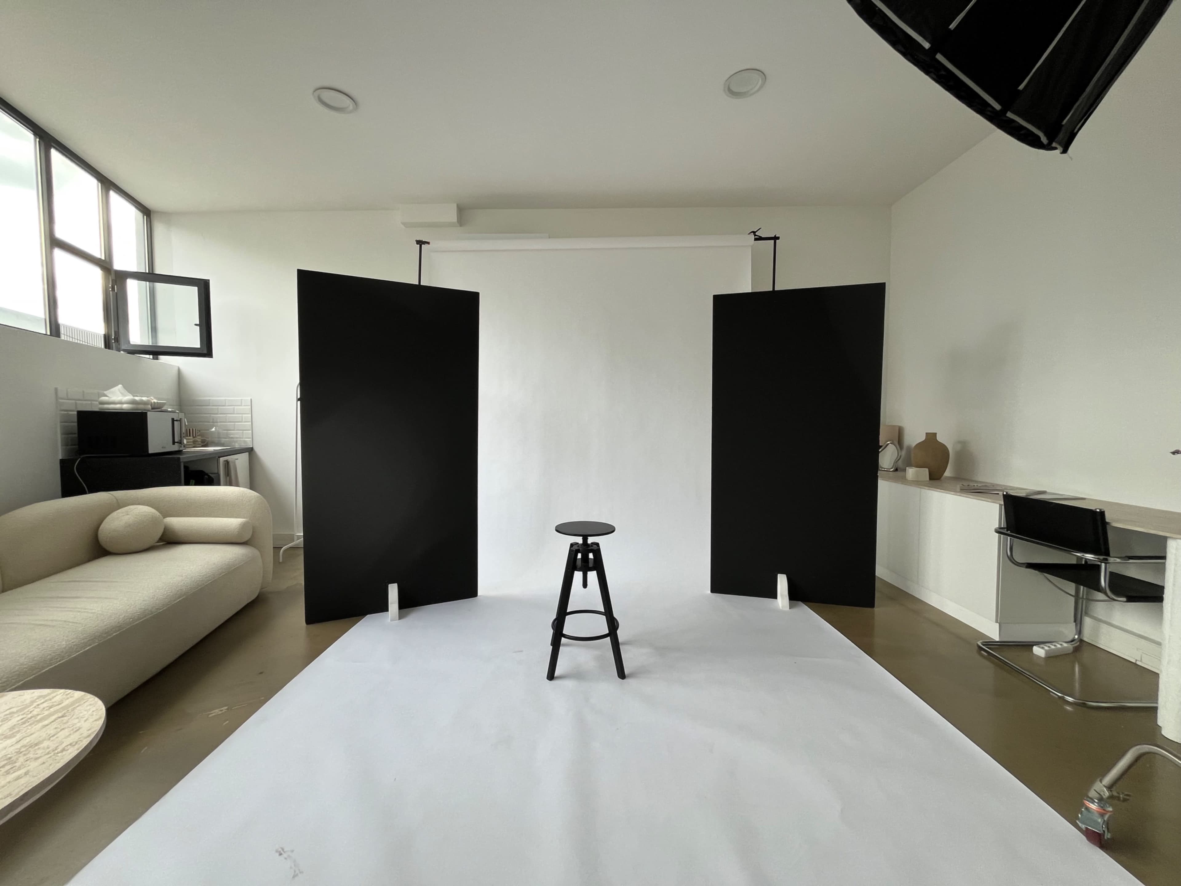 The image shows a photography studio with a black stool positioned on a white backdrop, flanked by two black panels, with a minimalist seating area and a desk in the background.