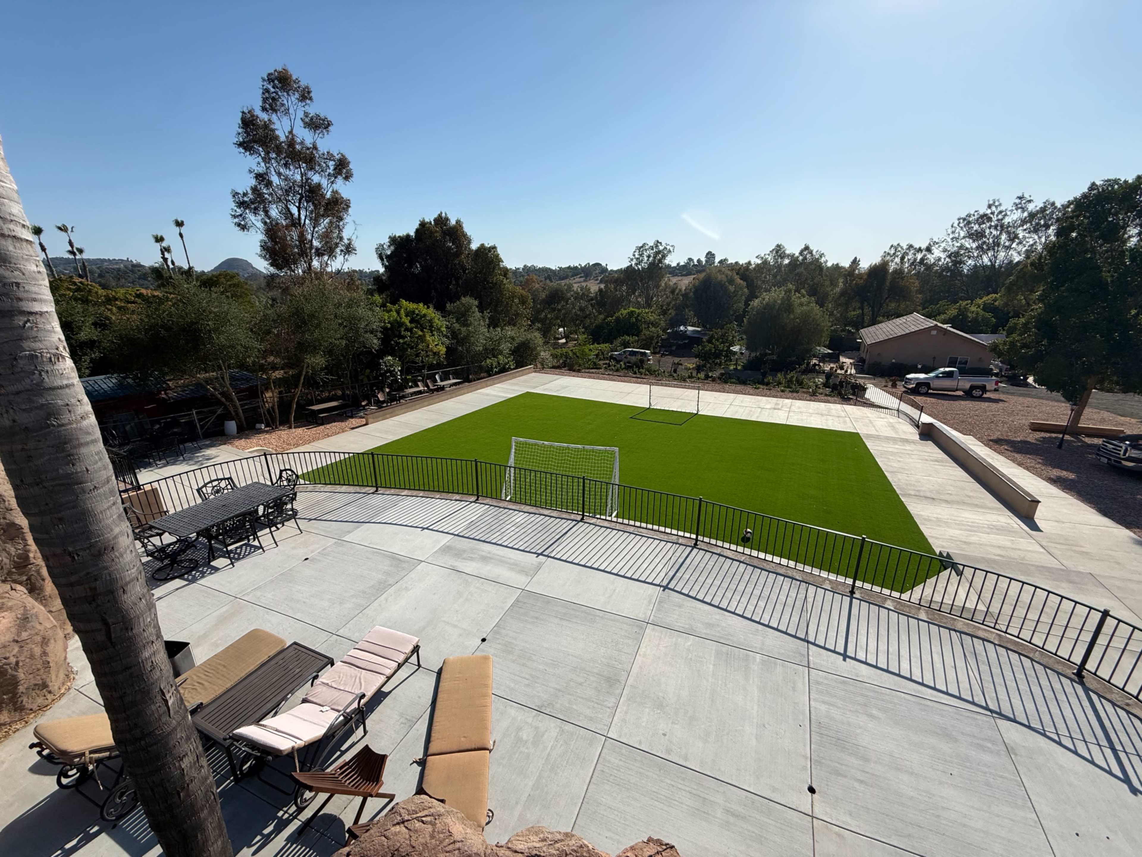 The image shows a large, green turf area with a soccer goal, surrounded by trees and a patio with lounge chairs and tables.