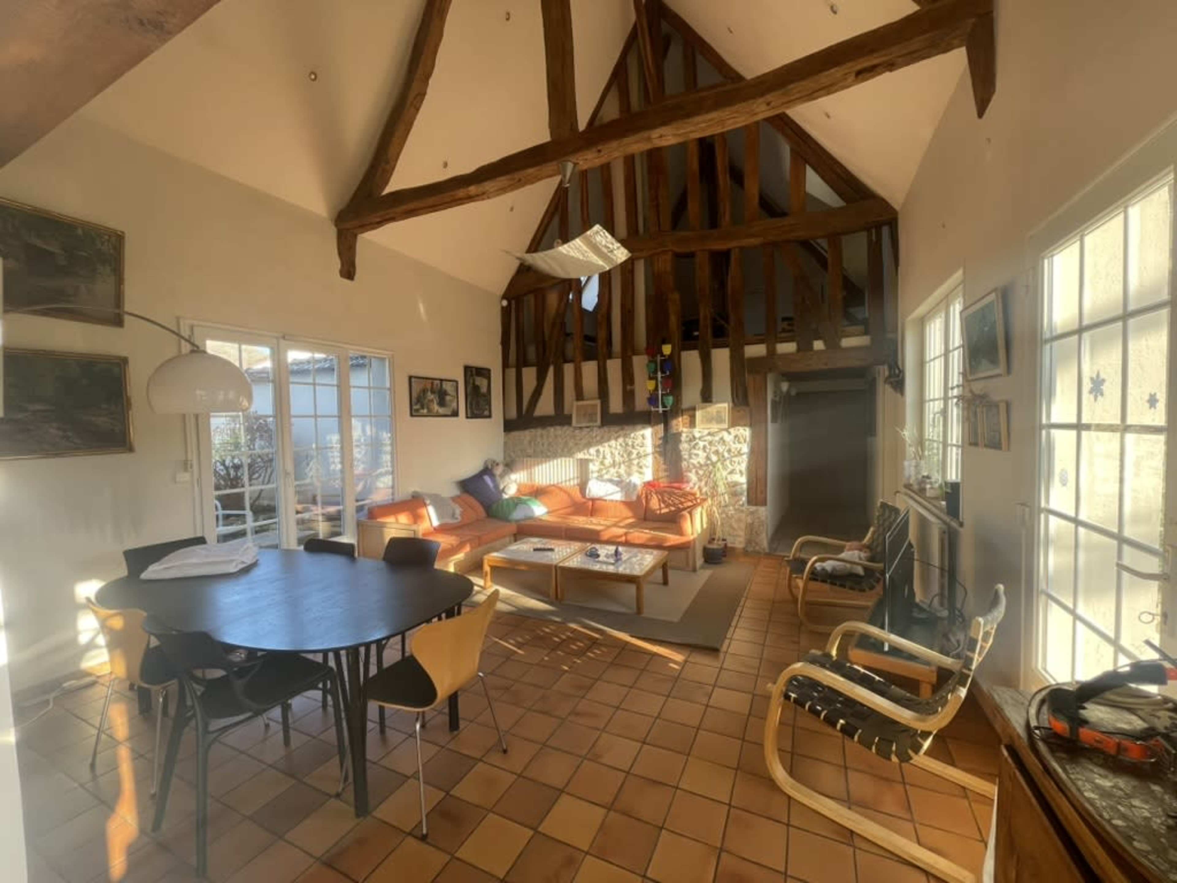 The image depicts a spacious living room with high wooden beams, a round dining table, a cozy seating area with orange couches, and large windows letting in natural light.