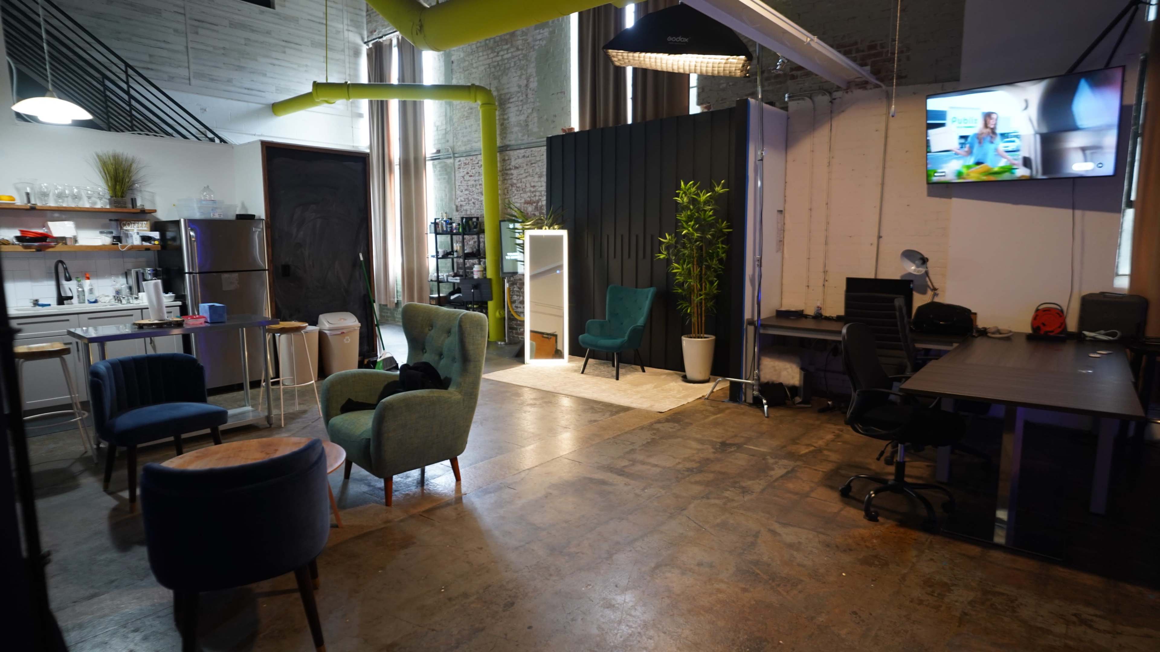The image shows an open workspace with a kitchen area, two green chairs, a dark sofa, and a desk, illuminated by natural light and overhead lamps.