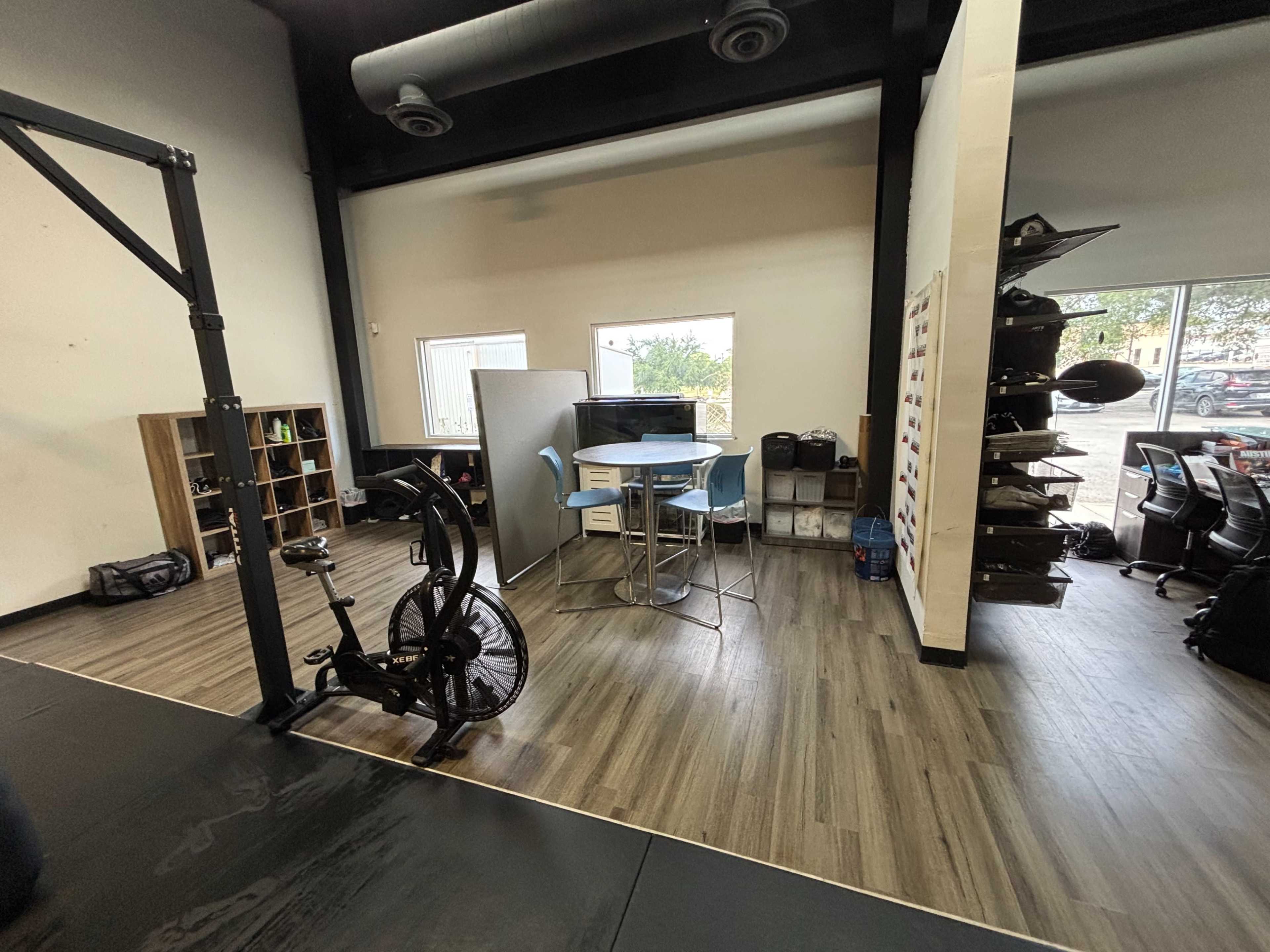 The image shows an interior space featuring exercise equipment, a table with chairs, and various storage units along the walls.