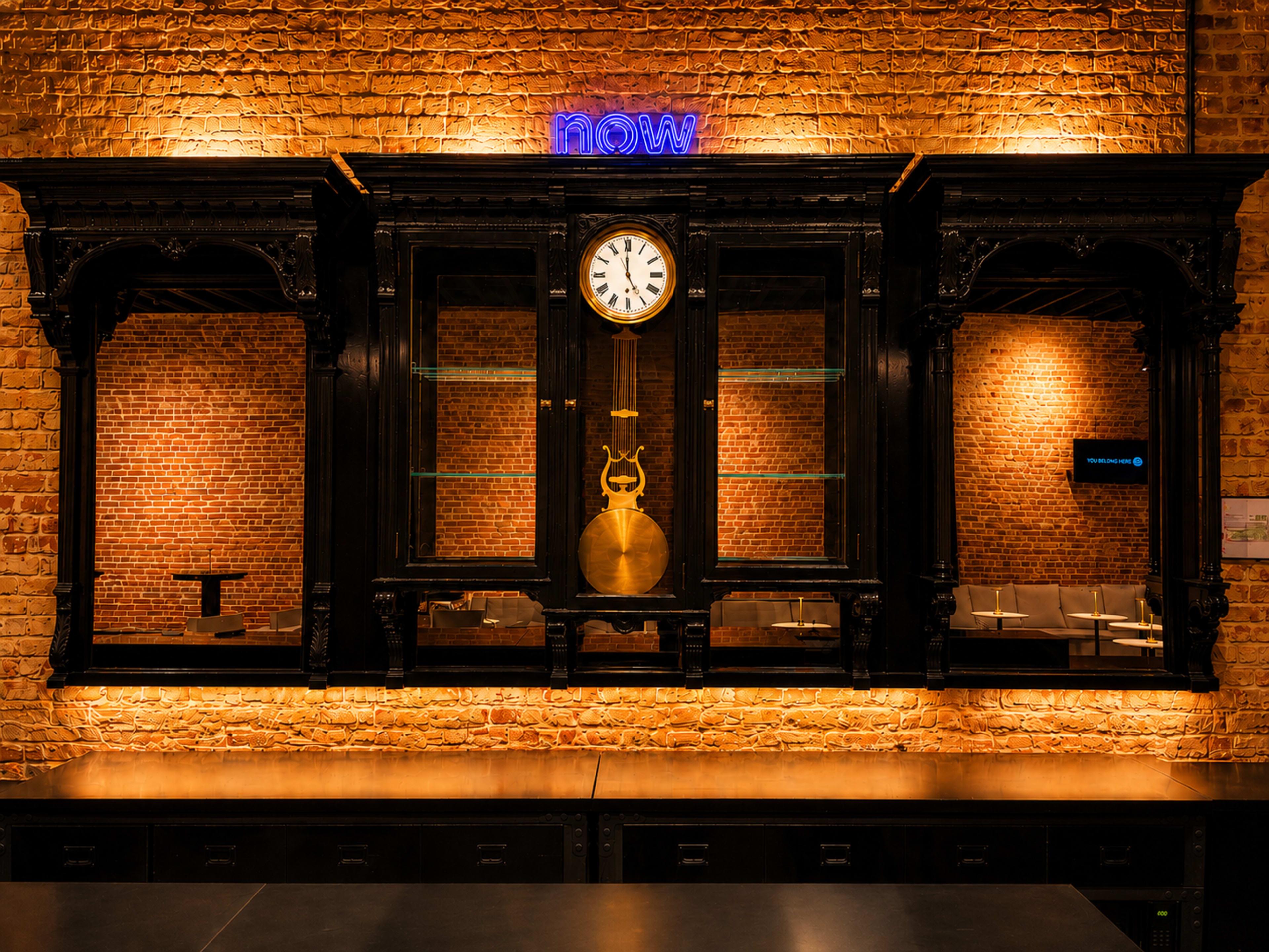 The image features a vintage black wooden display cabinet with a round clock and a brass bell, set against a brick wall illuminated by warm light and a neon sign that reads "now."