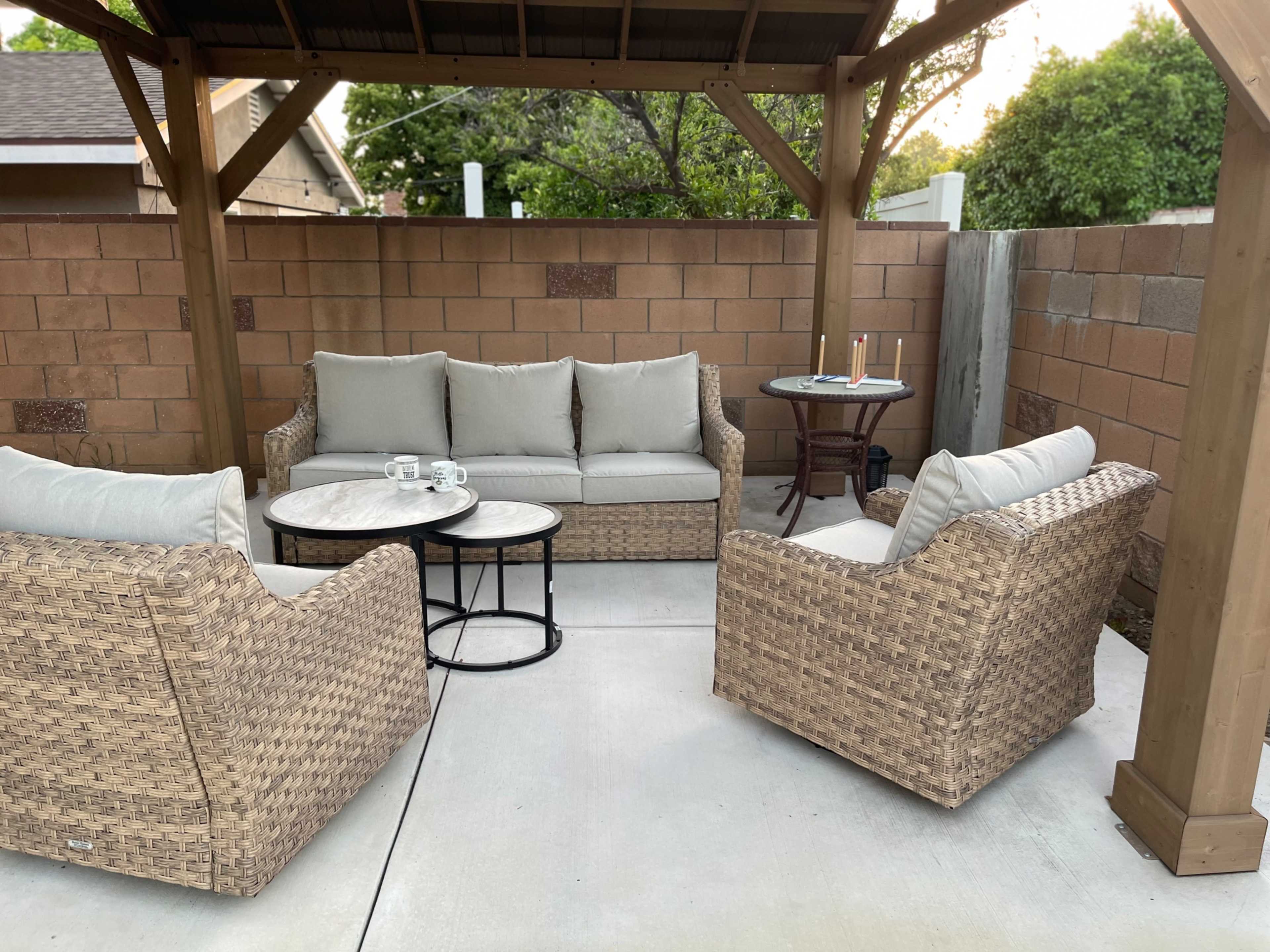 A patio area with a wicker sofa and armchairs arranged around a circular table under a wooden gazebo.