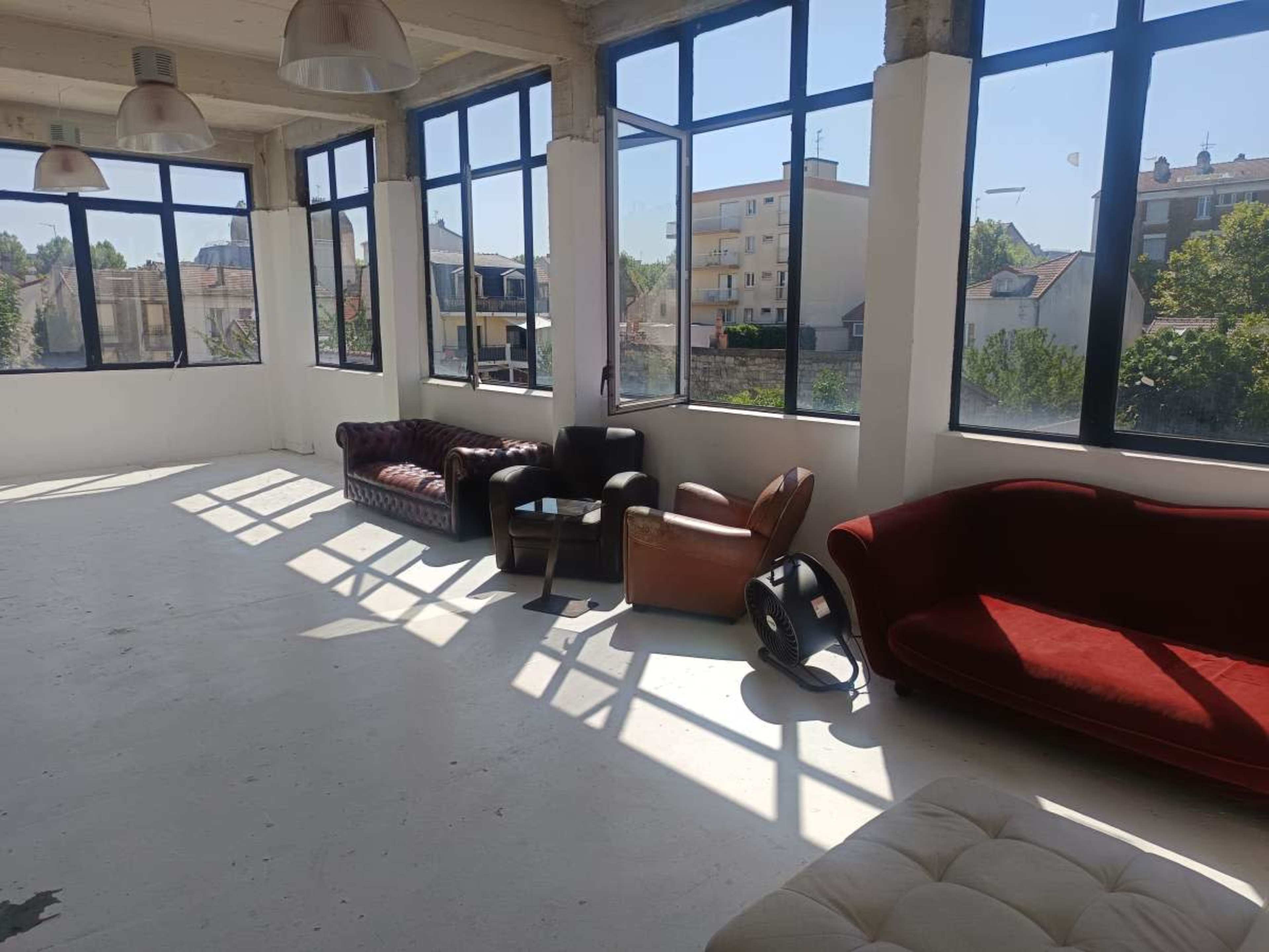 The image shows a brightly lit room with large windows, featuring several couches and armchairs arranged along one side.