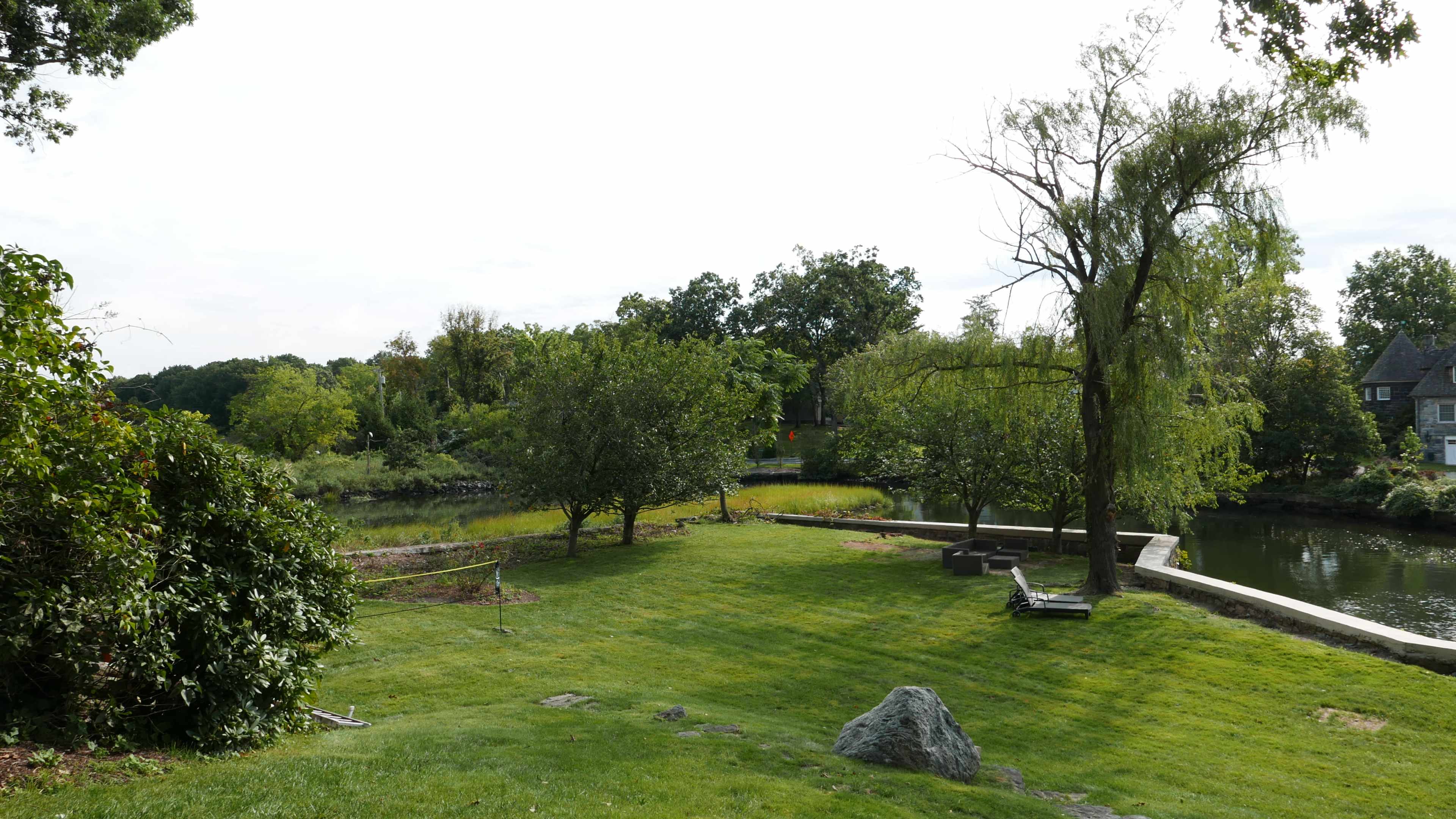 A grassy area features trees and a small pond in the background, with a stone and a bench positioned nearby.