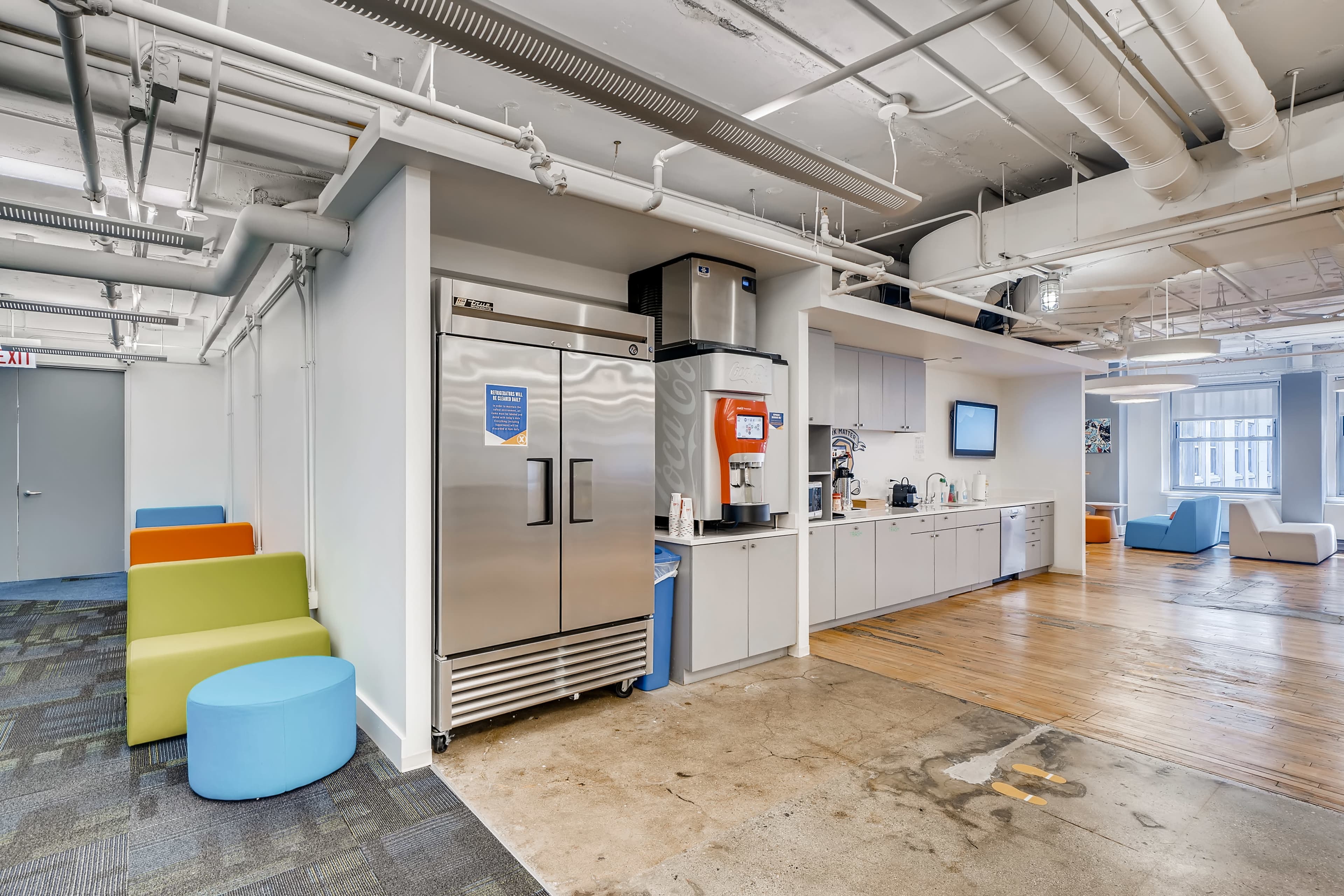 A modern office break room featuring a refrigerator, coffee machine, and seating areas with colorful chairs and tables.