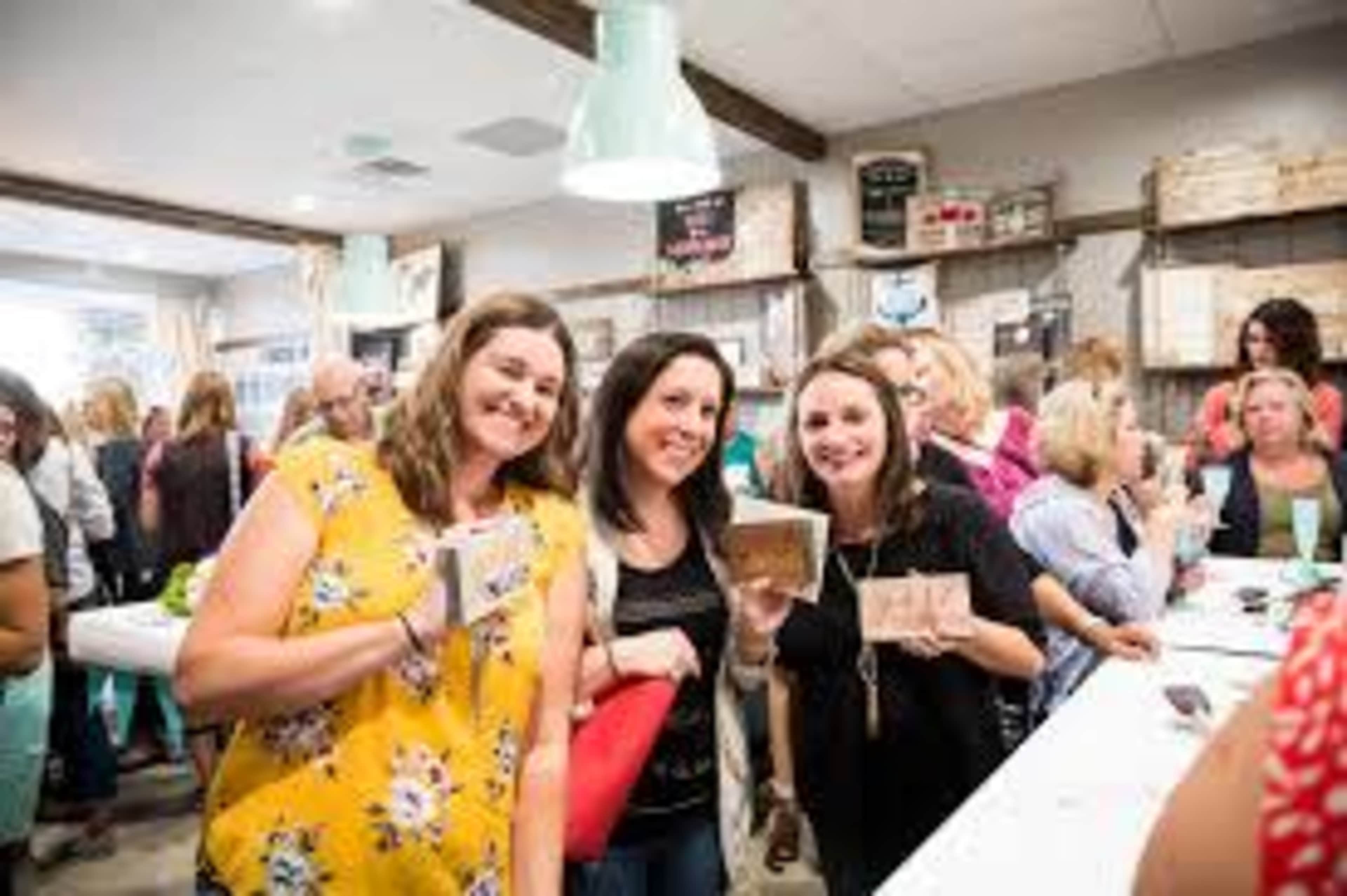 A group of three women pose with art projects in a busy workshop filled with people.