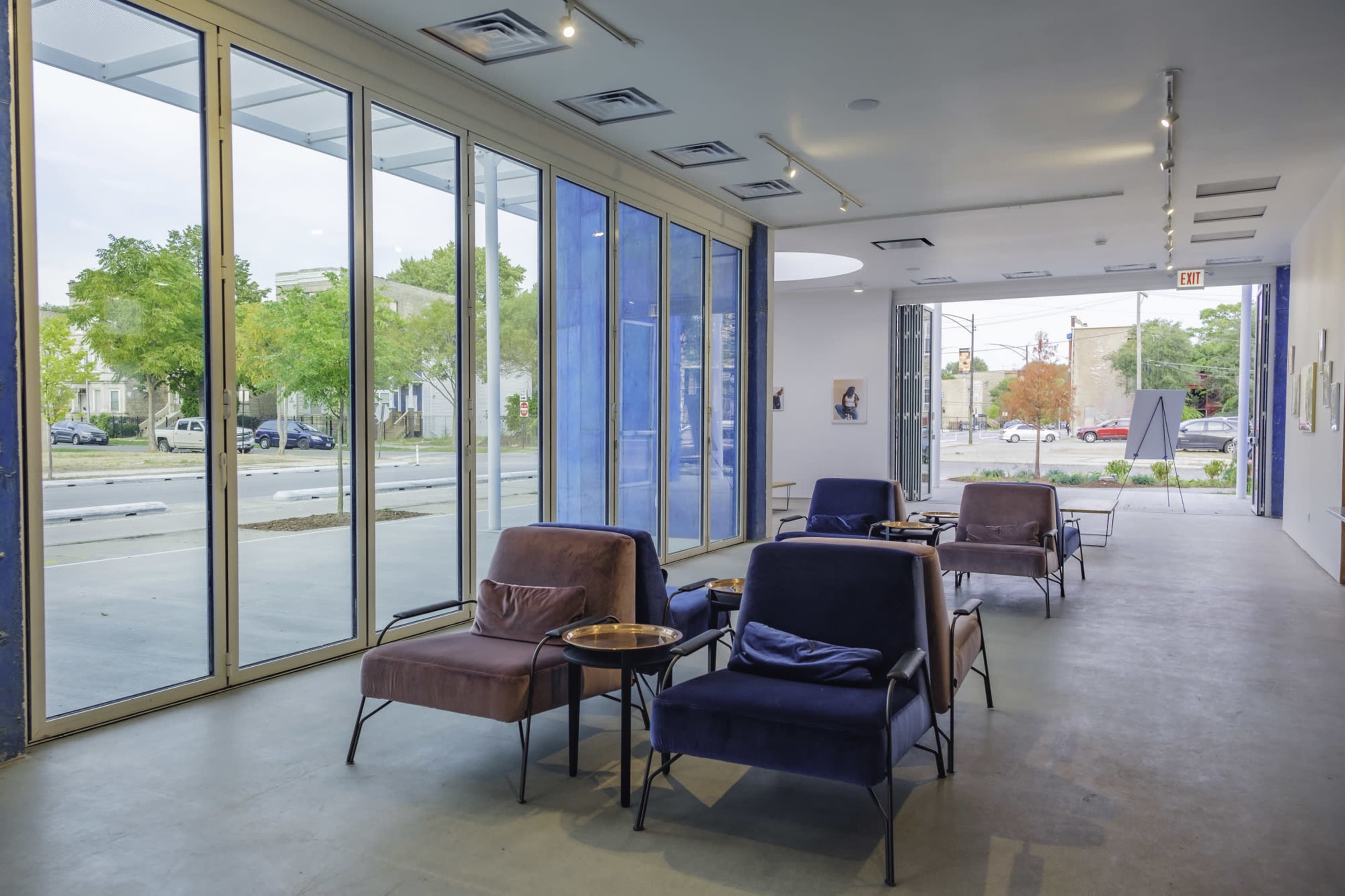 The image shows a modern interior space featuring several comfortable chairs arranged around small tables, with large glass windows allowing natural light to enter.