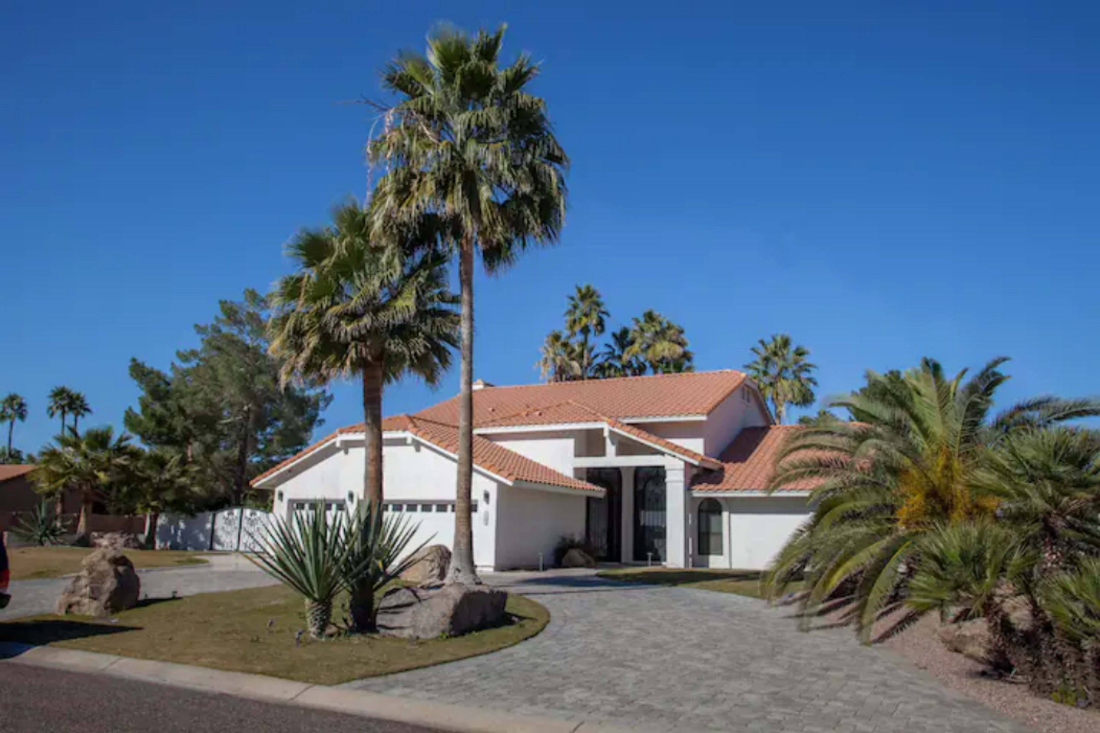 A modern house with a red-tiled roof is surrounded by palm trees and sits on a paved driveway.
