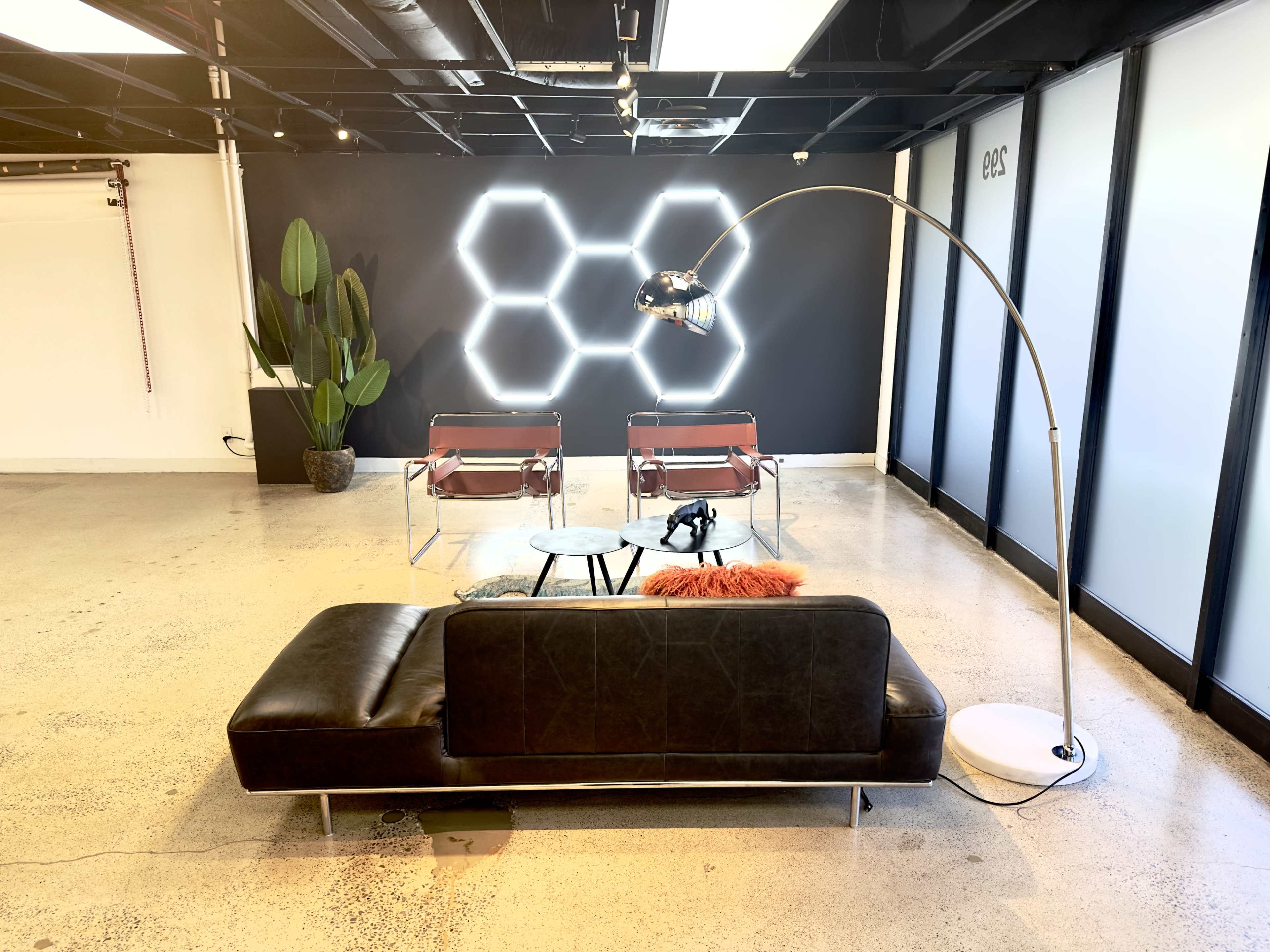The image shows a modern lounge area featuring a dark sofa, two chairs, a small coffee table, and a decorative plant against a geometric neon light backdrop.