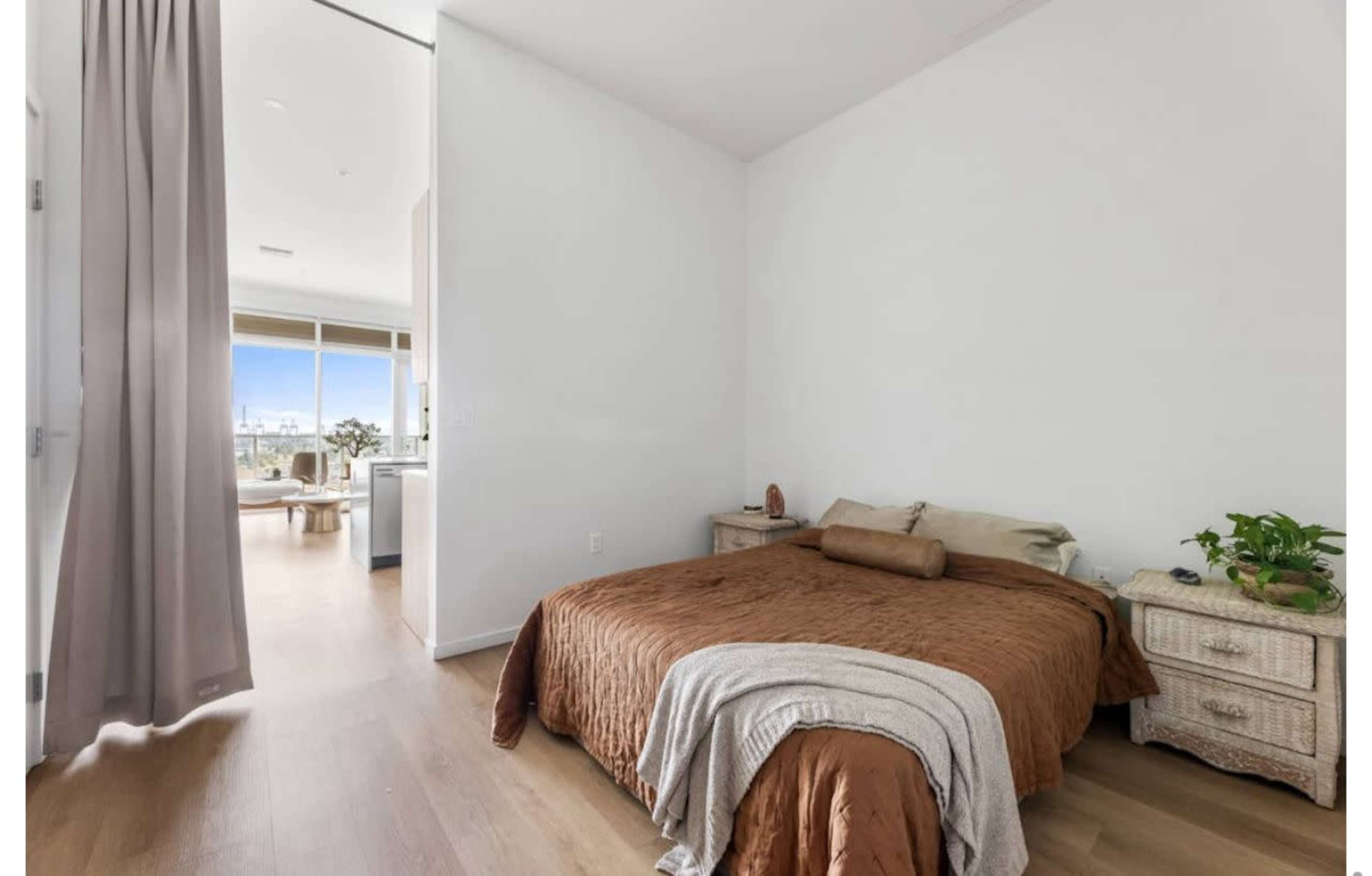 The image shows a bedroom with a brown bedspread, a bedside table with plants, and a large window revealing a view outside.