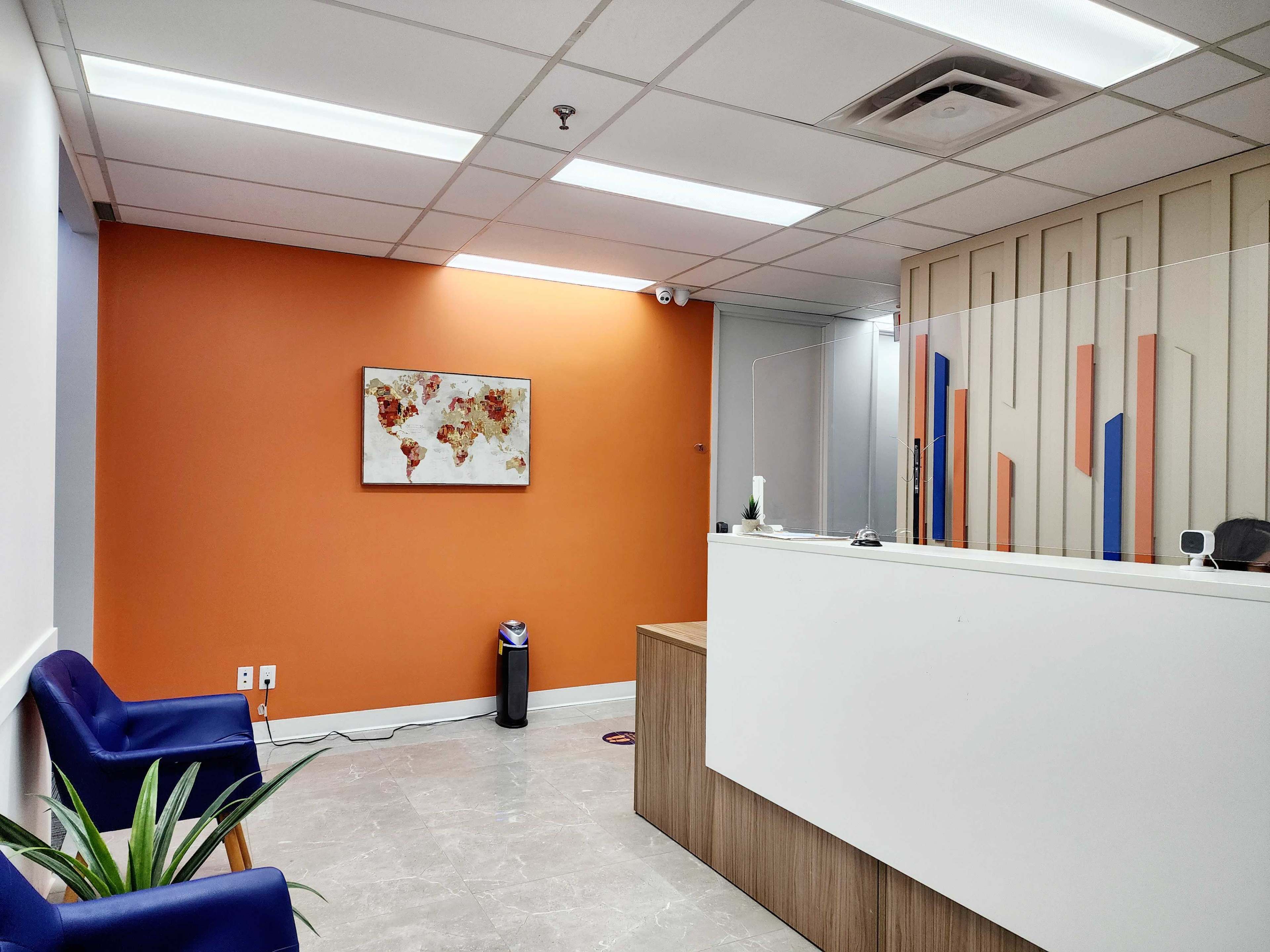 A reception area with an orange accent wall, a world map artwork, a white front desk, blue seating, and a plant in the corner.