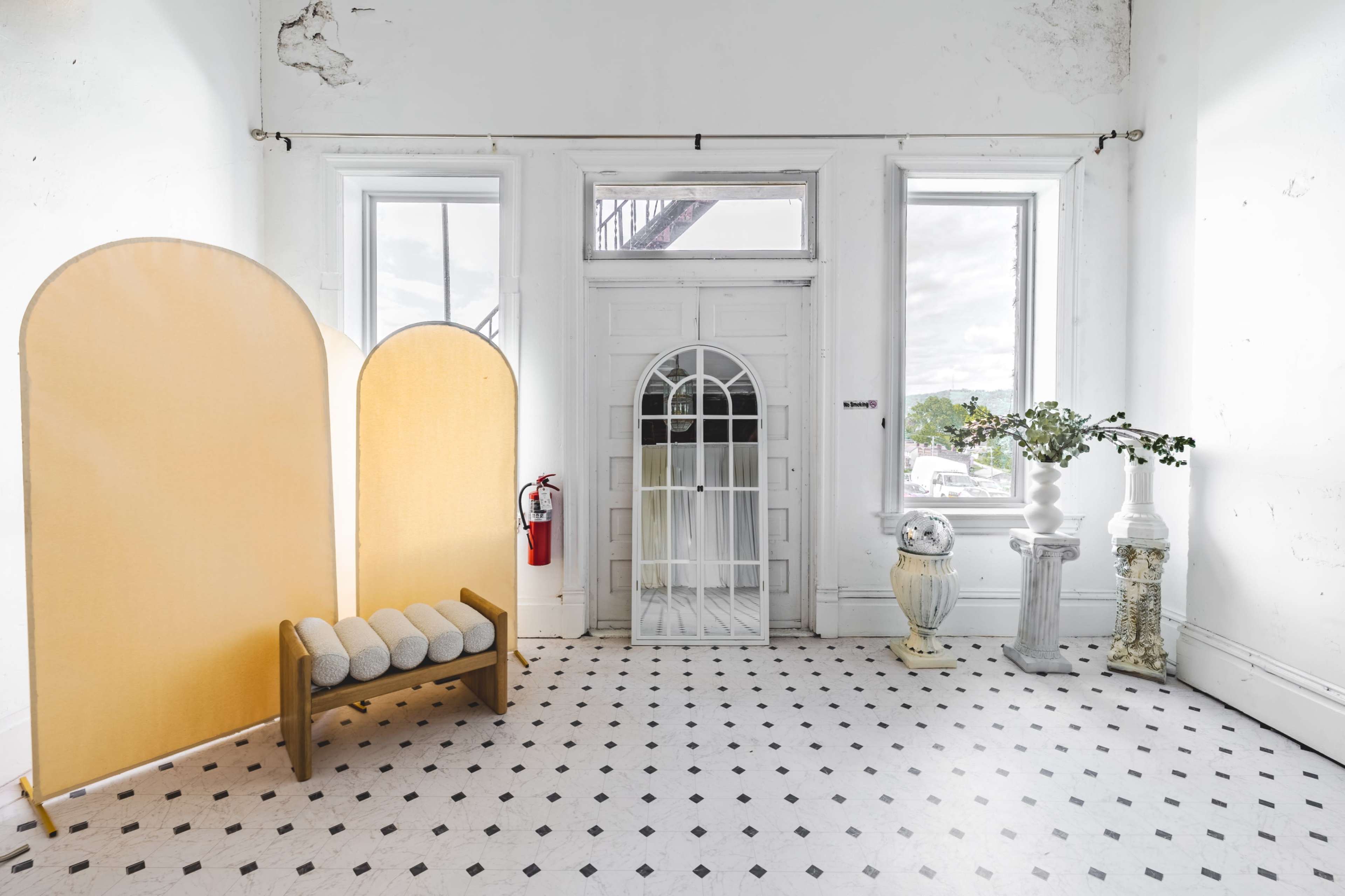 A bright, minimalist interior featuring a wooden bench, two tall yellow panels, and decorative columns flanking a white door with arched glass.