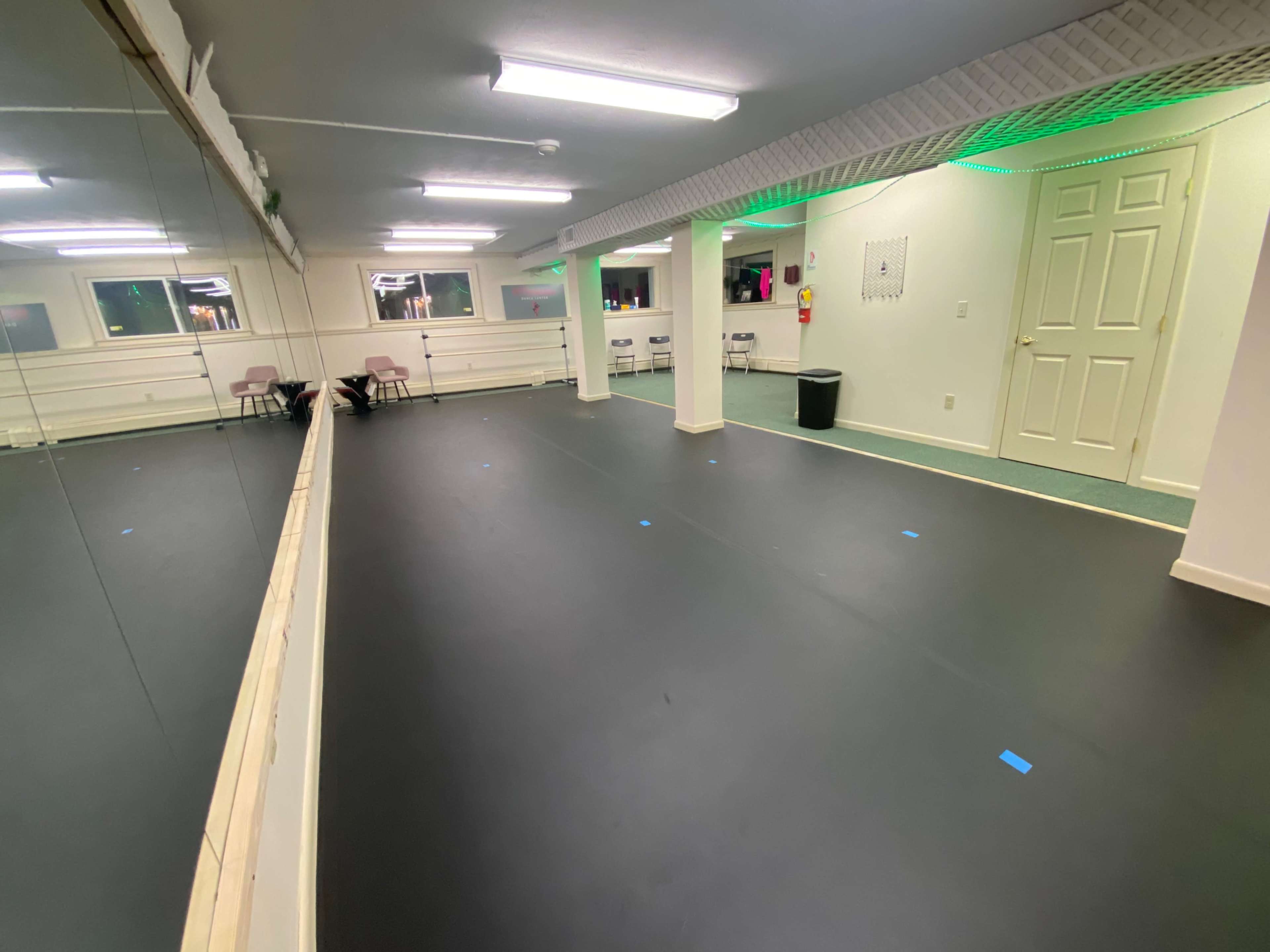 A spacious room with black flooring, mirrored walls, and subtle green lighting, featuring a few chairs and a trash can.