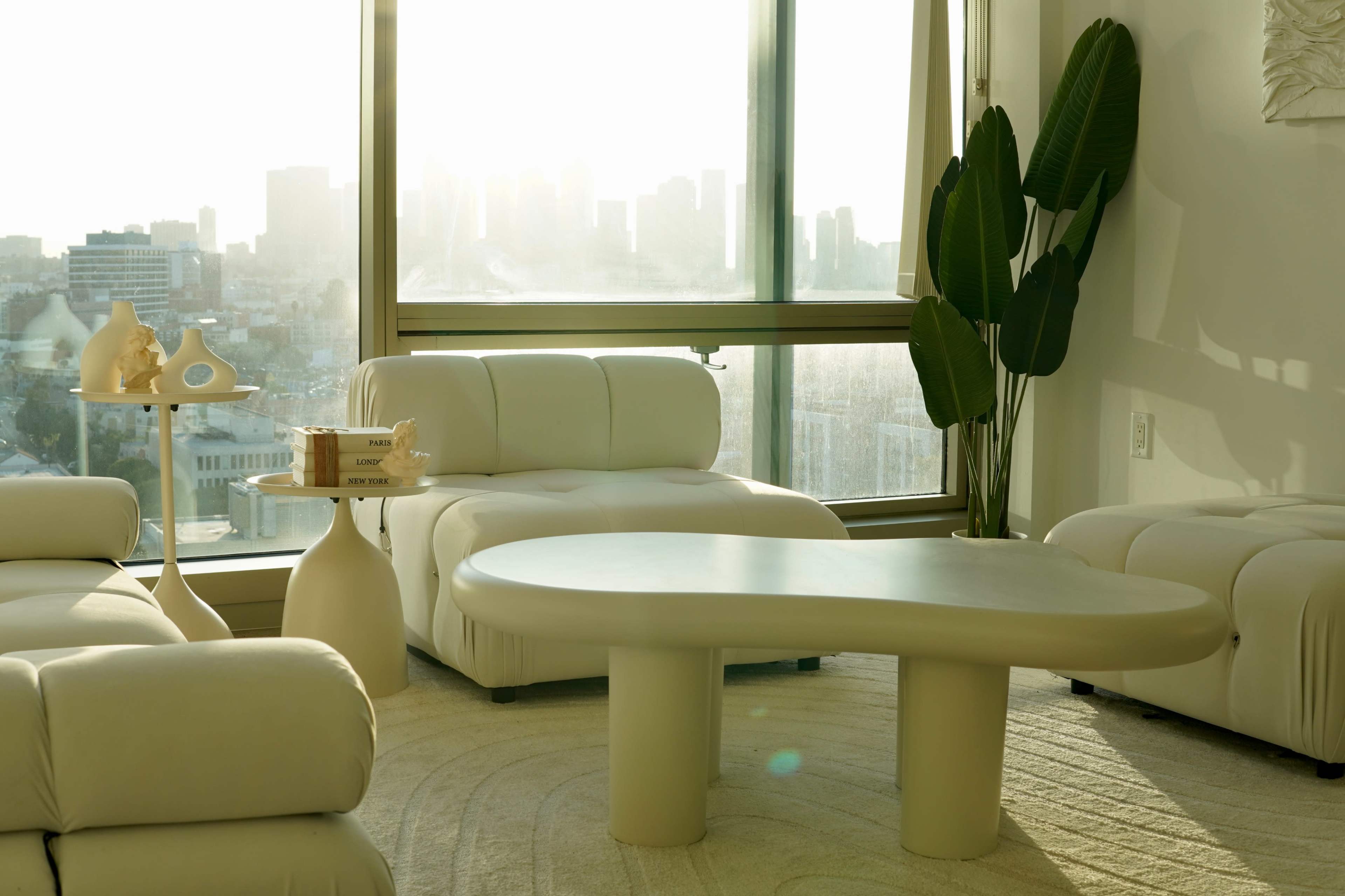 A modern living room features a curved beige coffee table, two white sofas, and a large window with a cityscape view.