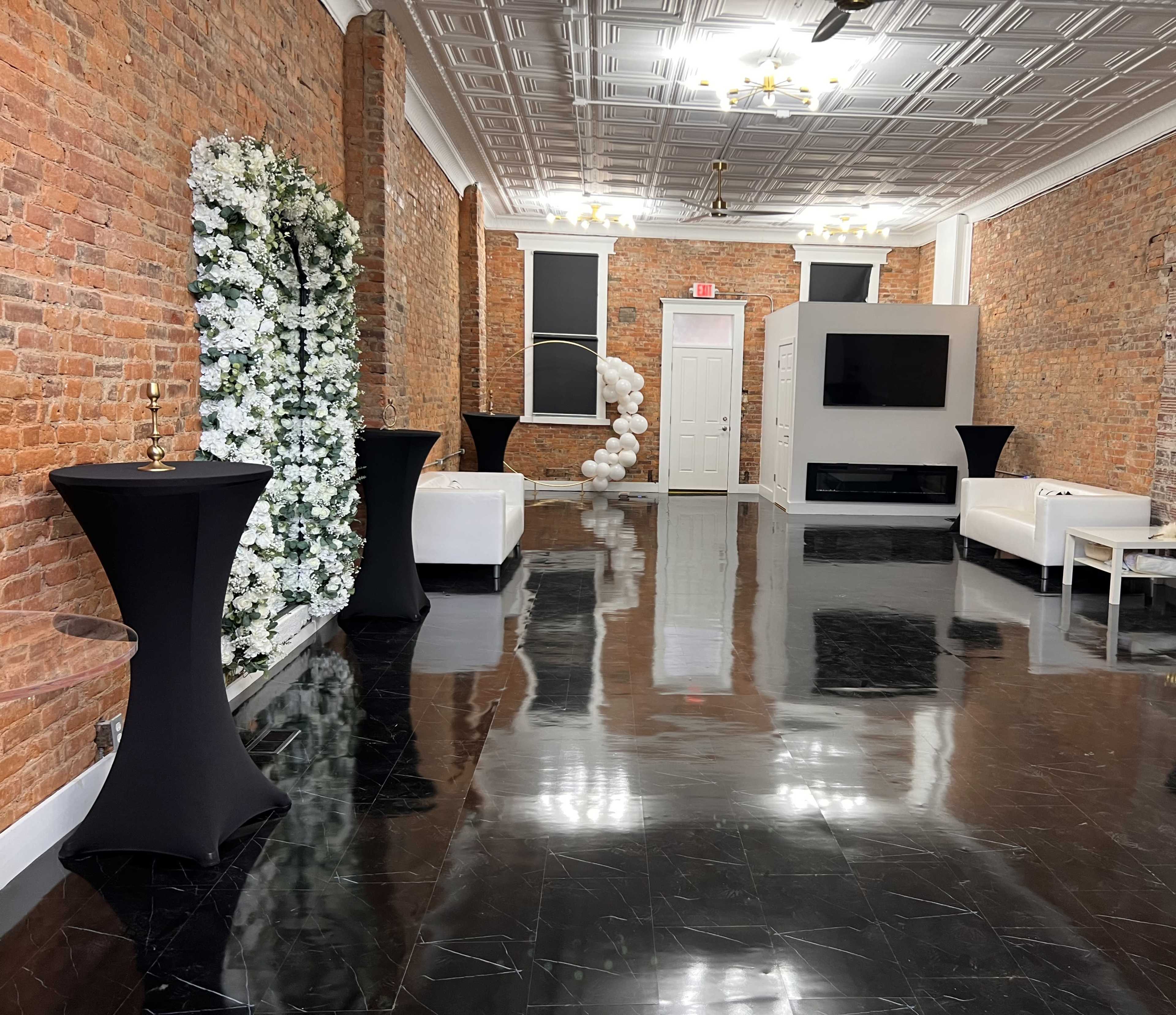 The image shows a decorated interior space with exposed brick walls, black floor tiles, two white sofas, and black cocktail tables.