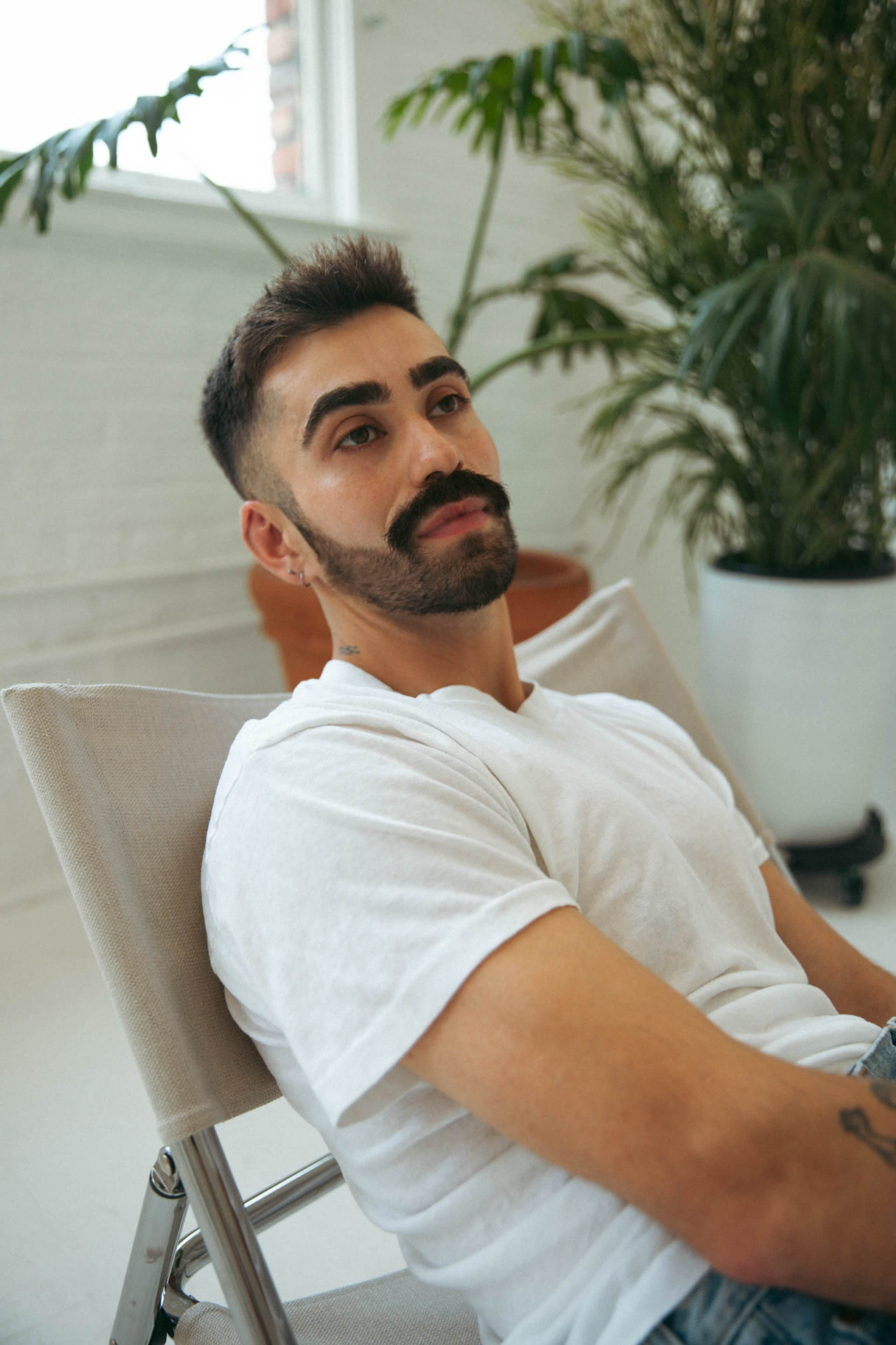 A man with a mustache sits relaxed in a chair, surrounded by plants in a bright, airy room.