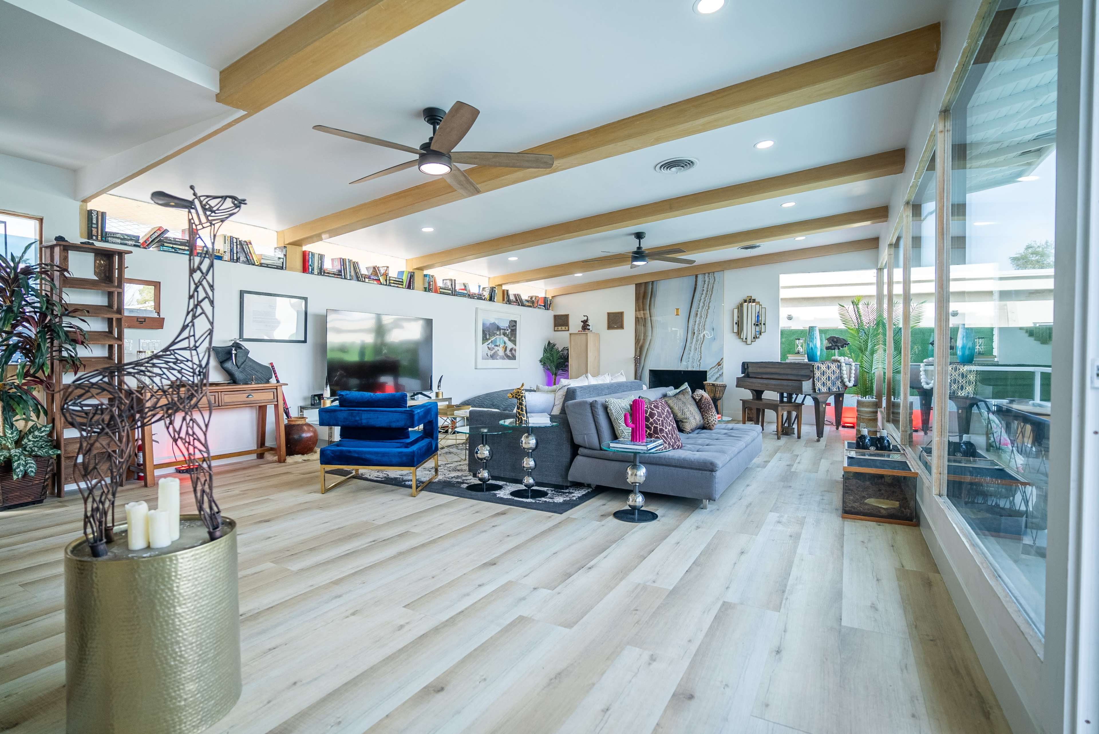 The image shows a modern living room with wooden beams, a mix of seating options, large windows, and decorative plants.