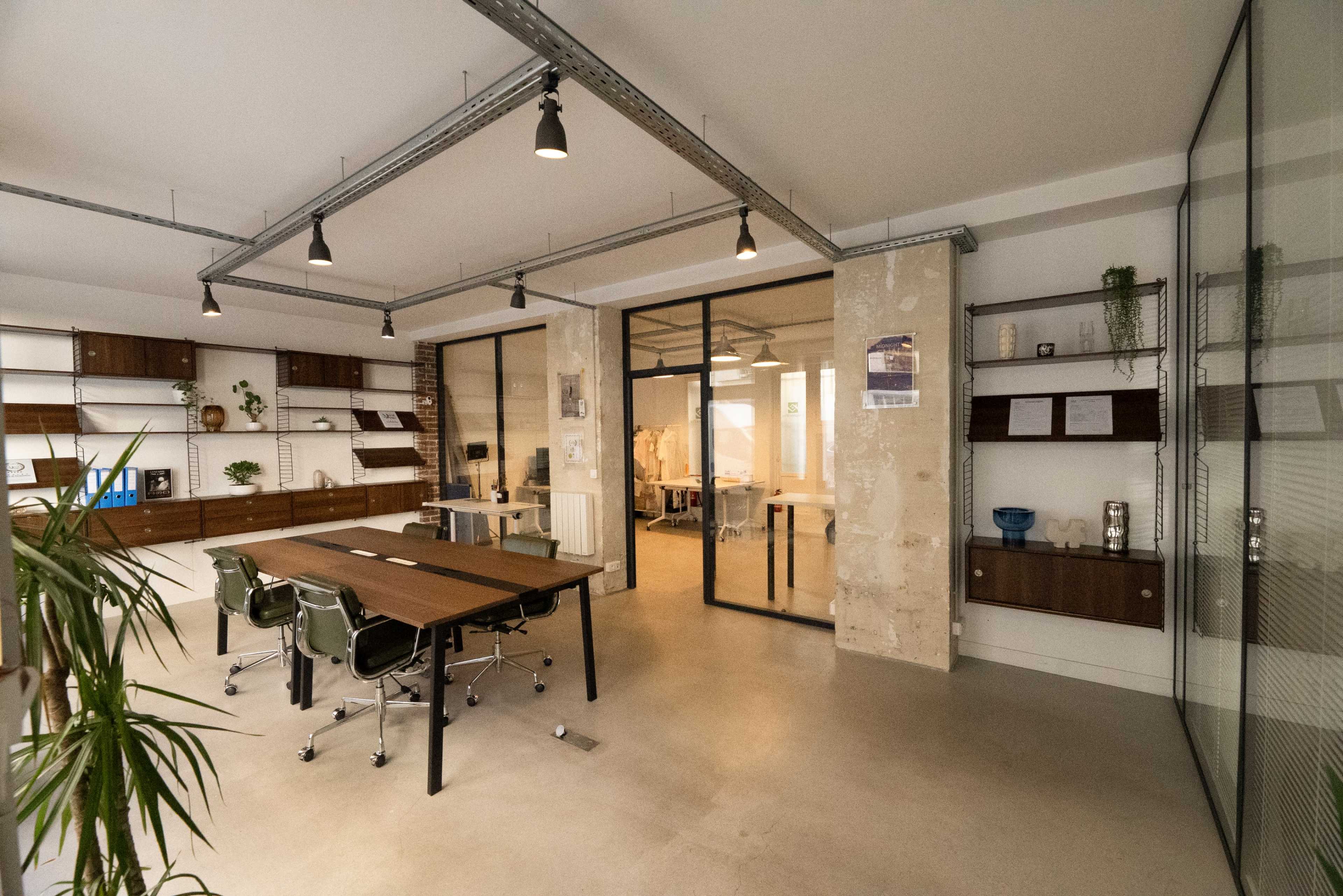 The image depicts a modern office space featuring a large wooden table surrounded by chairs, glass partitions, and shelves with decor and storage.