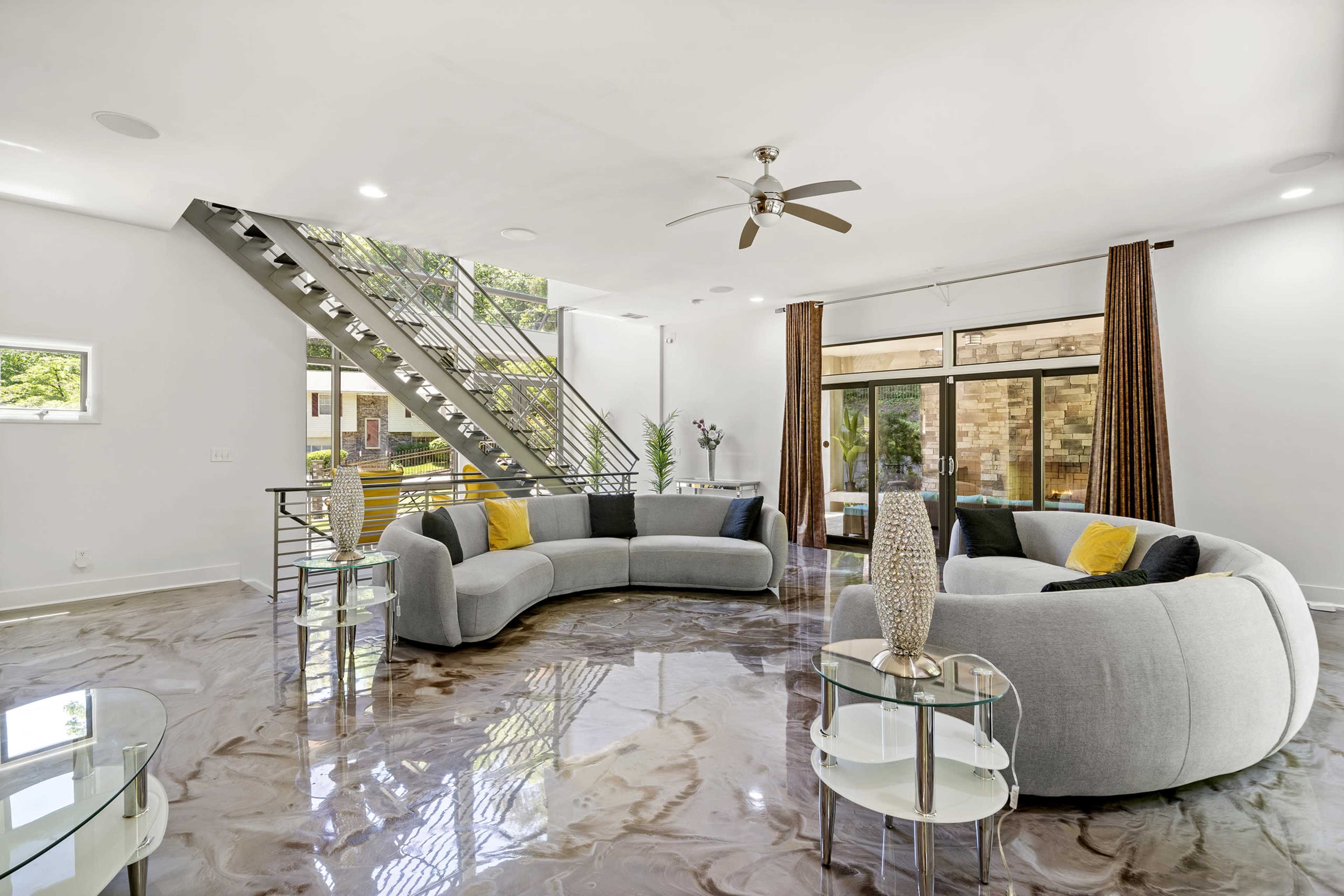 The living room features a curved sectional sofa, glass tables, and large windows overlooking a staircase with metal railings.