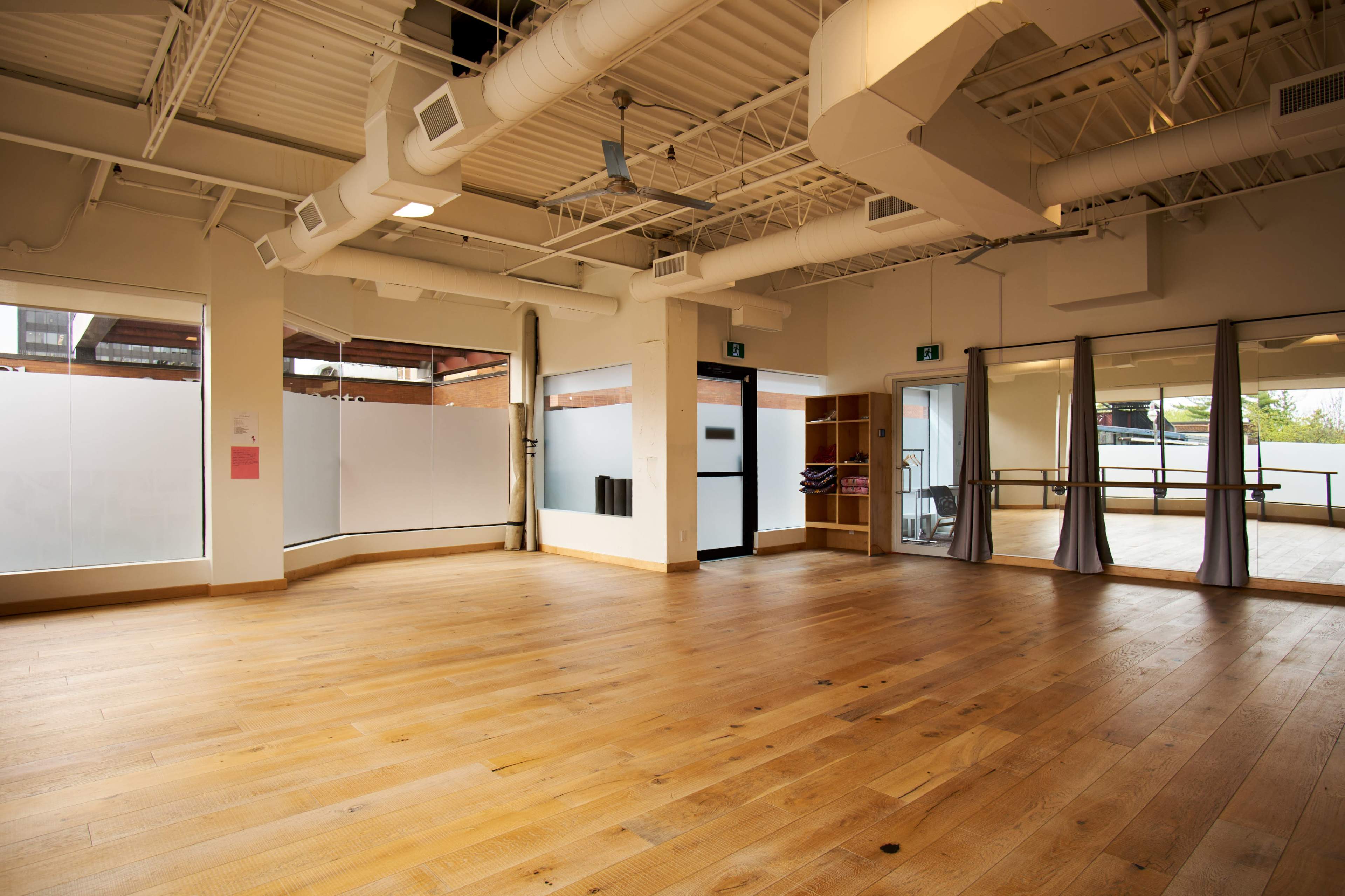 The image shows a spacious, empty studio with wooden flooring, large mirrors, and natural light streaming in through the windows.