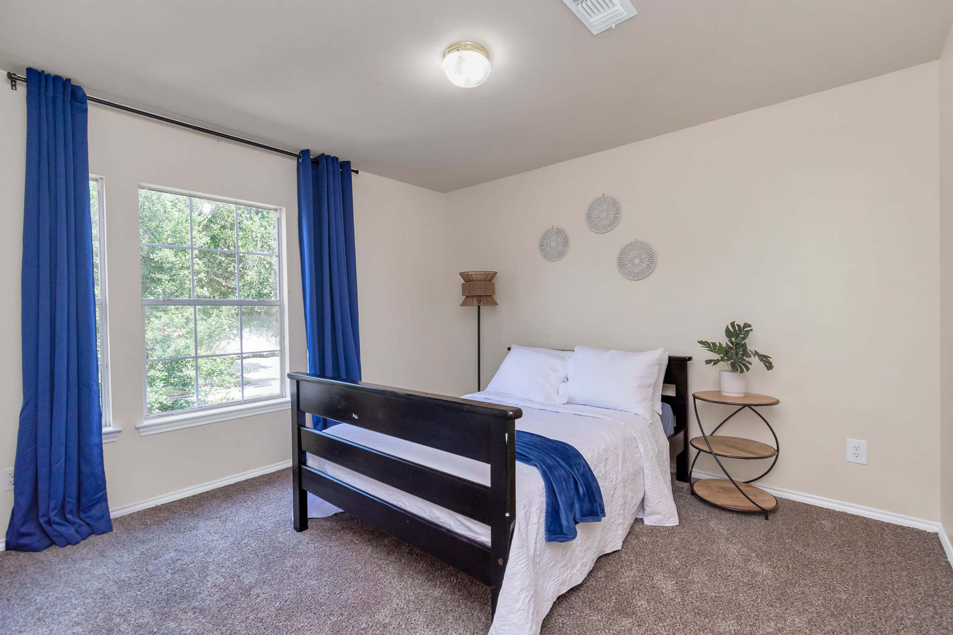 A simple bedroom features a bed with white linens, a round bedside table, and blue curtains framing a window.