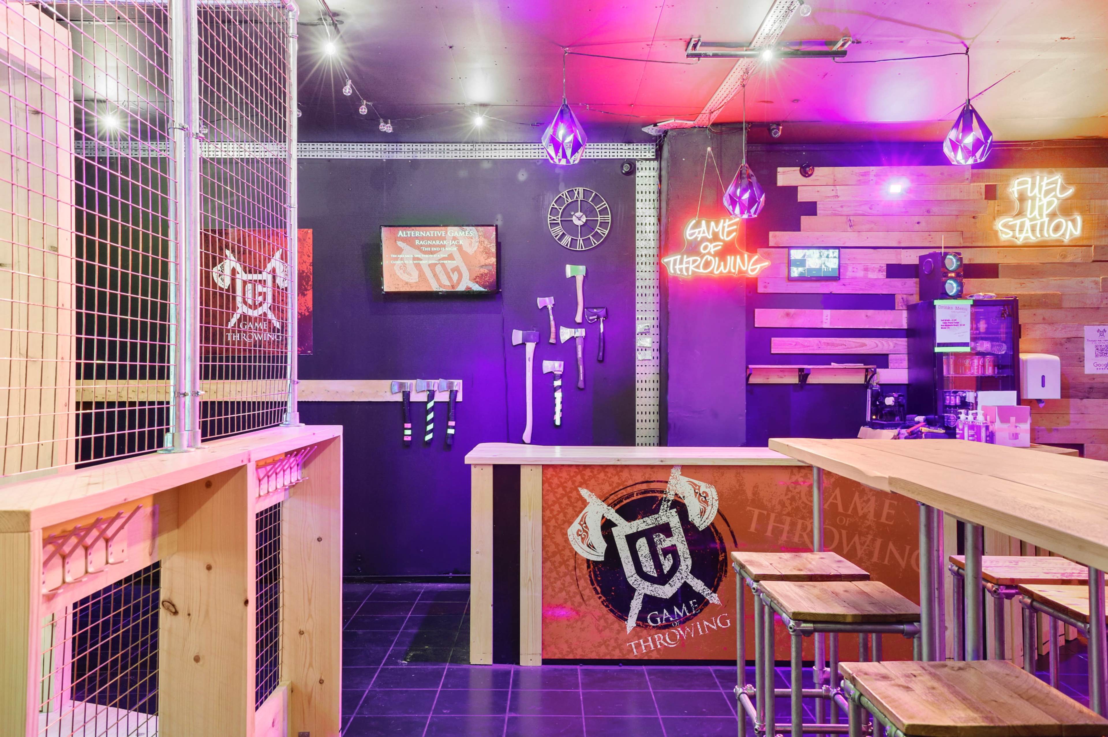 An indoor axe-throwing venue with a reception area featuring a wooden counter, neon signs, and an axe-throwing target against a dark wall.
