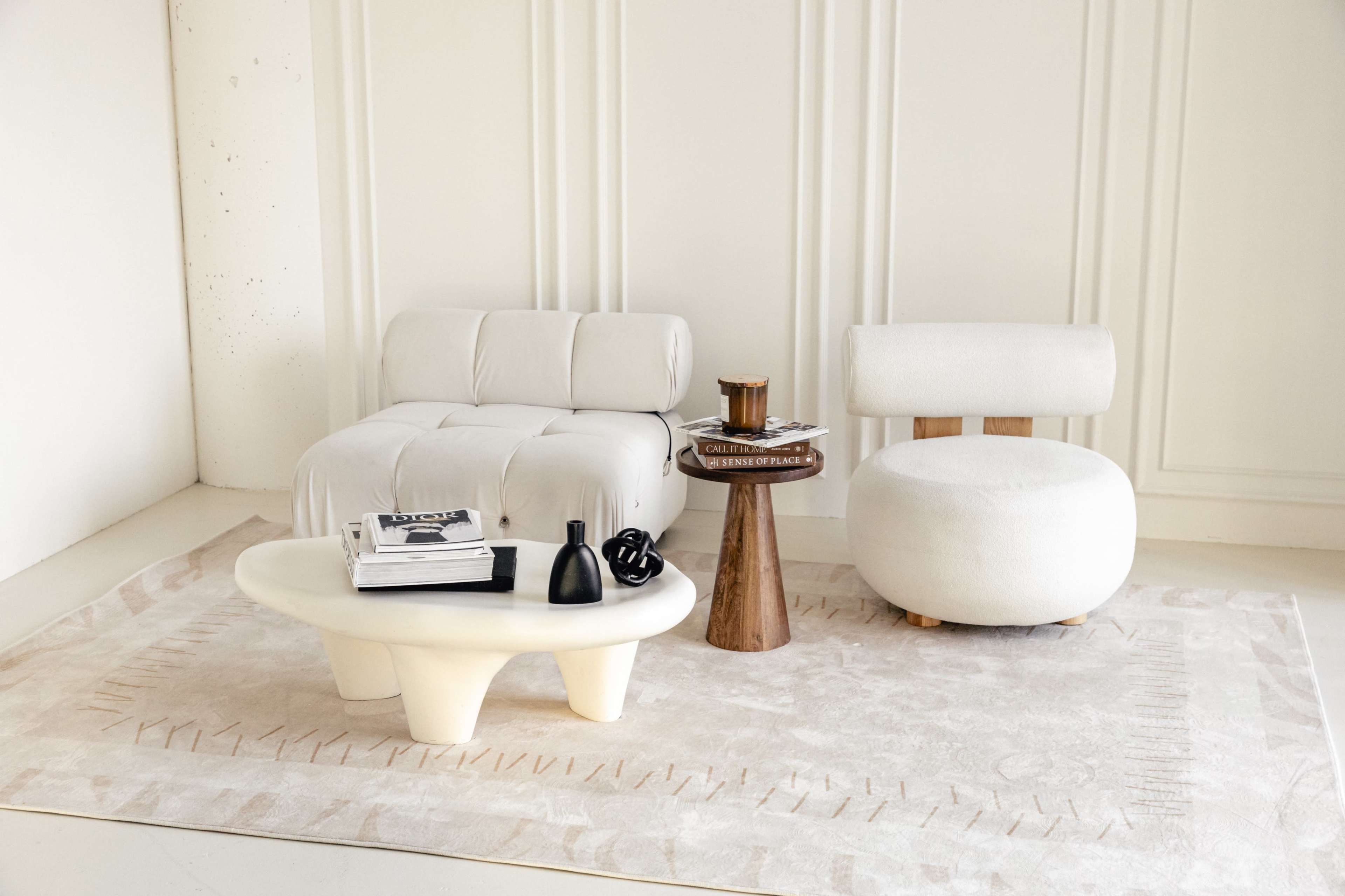The image displays a modern living room setup featuring a white, upholstered chaise lounge, a rounded armchair, a sculptural coffee table, and a small wooden side table on a light rug.