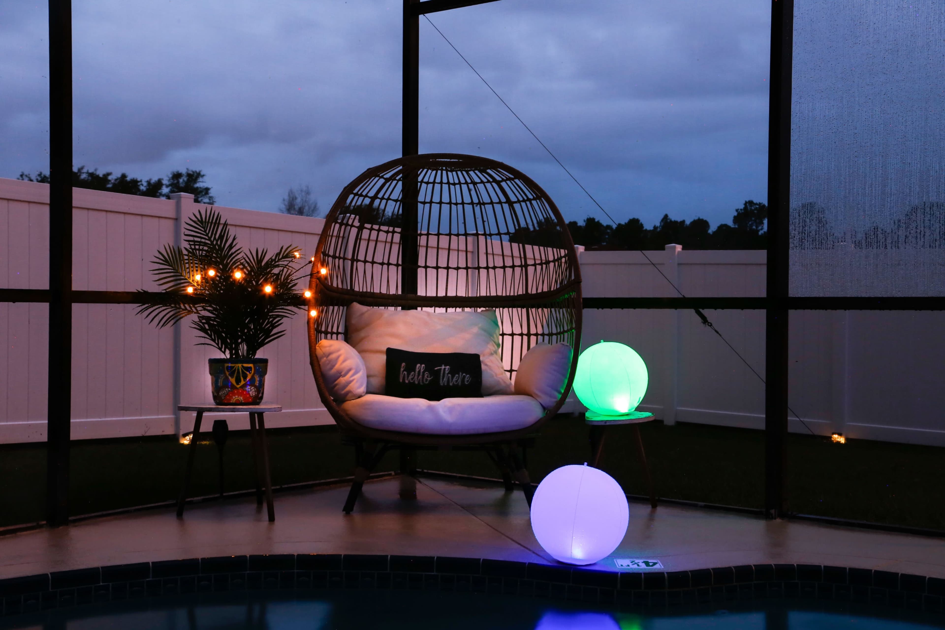 A hanging chair with cushions is situated beside a small table and illuminated orbs, set against a backdrop of a dimly lit outdoor area with a fence.
