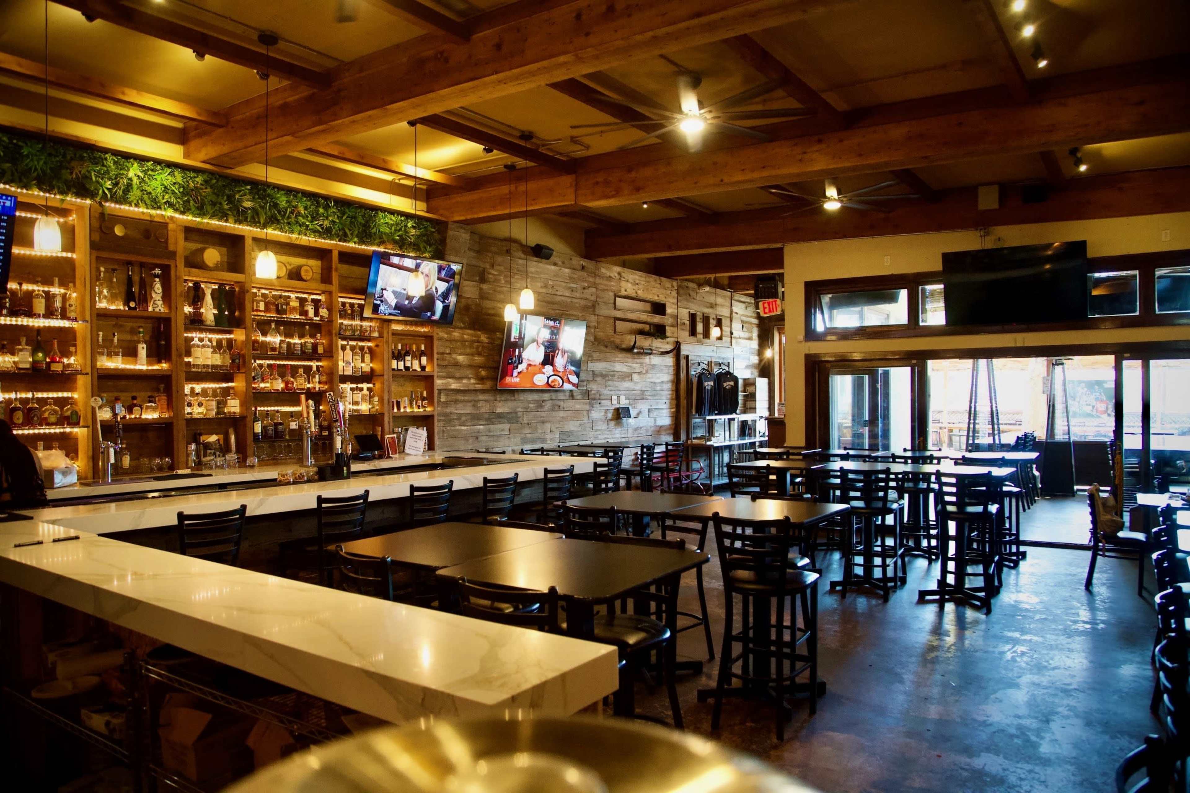 The image shows an interior view of a restaurant with wooden beams, a bar area stocked with bottles, and several tables arranged for dining.