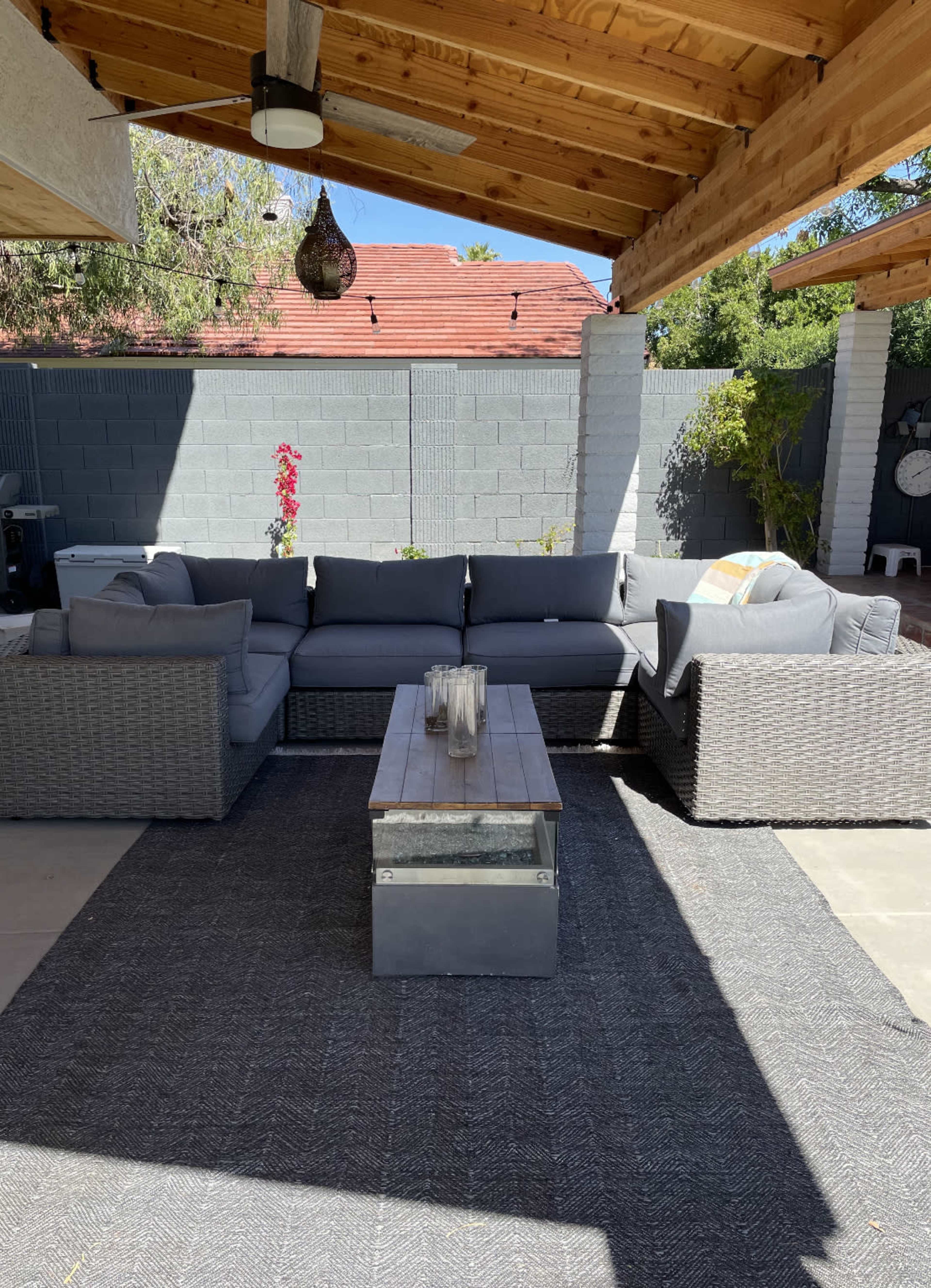 A modern outdoor seating area features a gray wicker sectional sofa, a wooden coffee table, and a patterned rug, all situated under a wooden pergola.