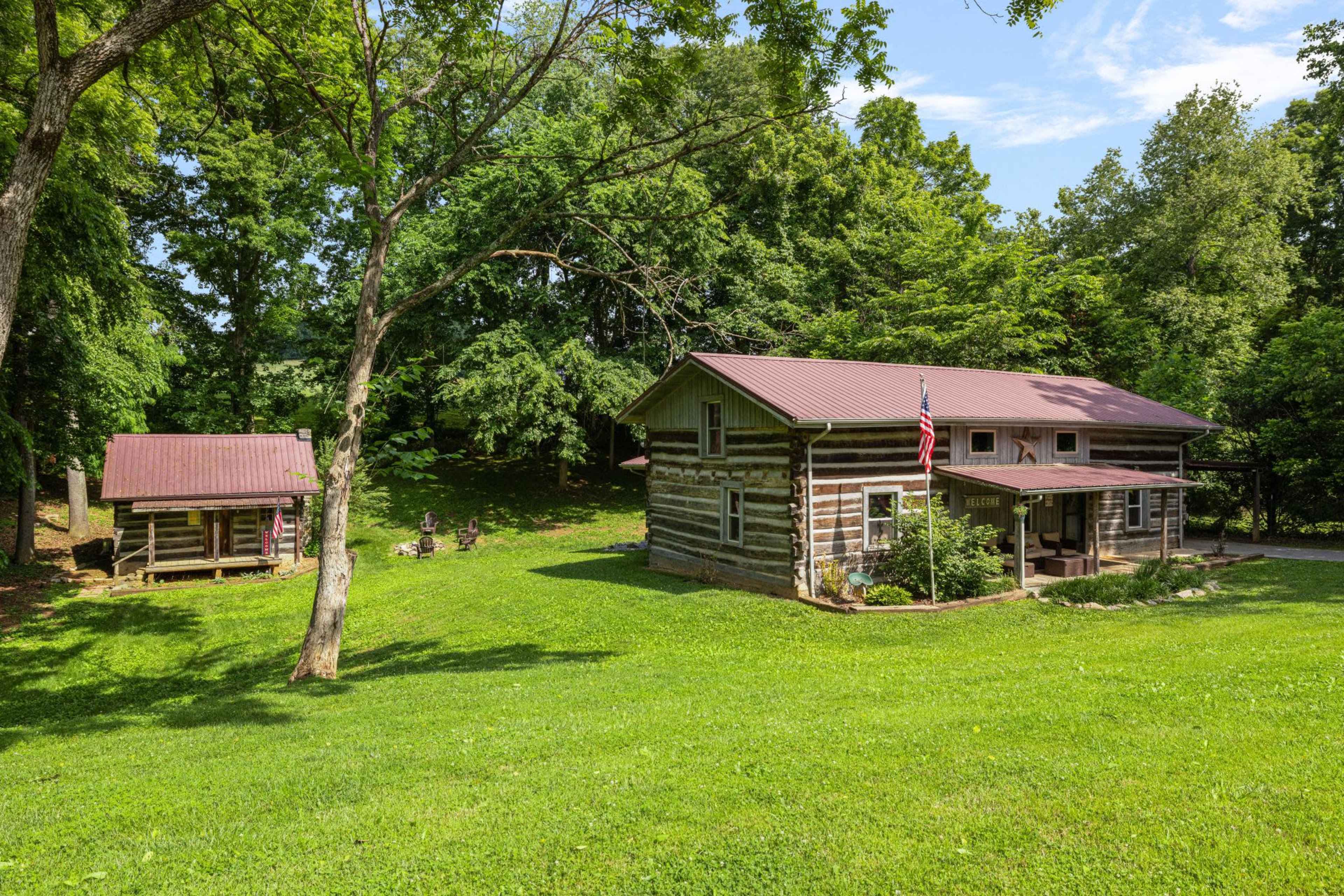 The image shows a log cabin and a smaller building in a lush green landscape surrounded by trees.