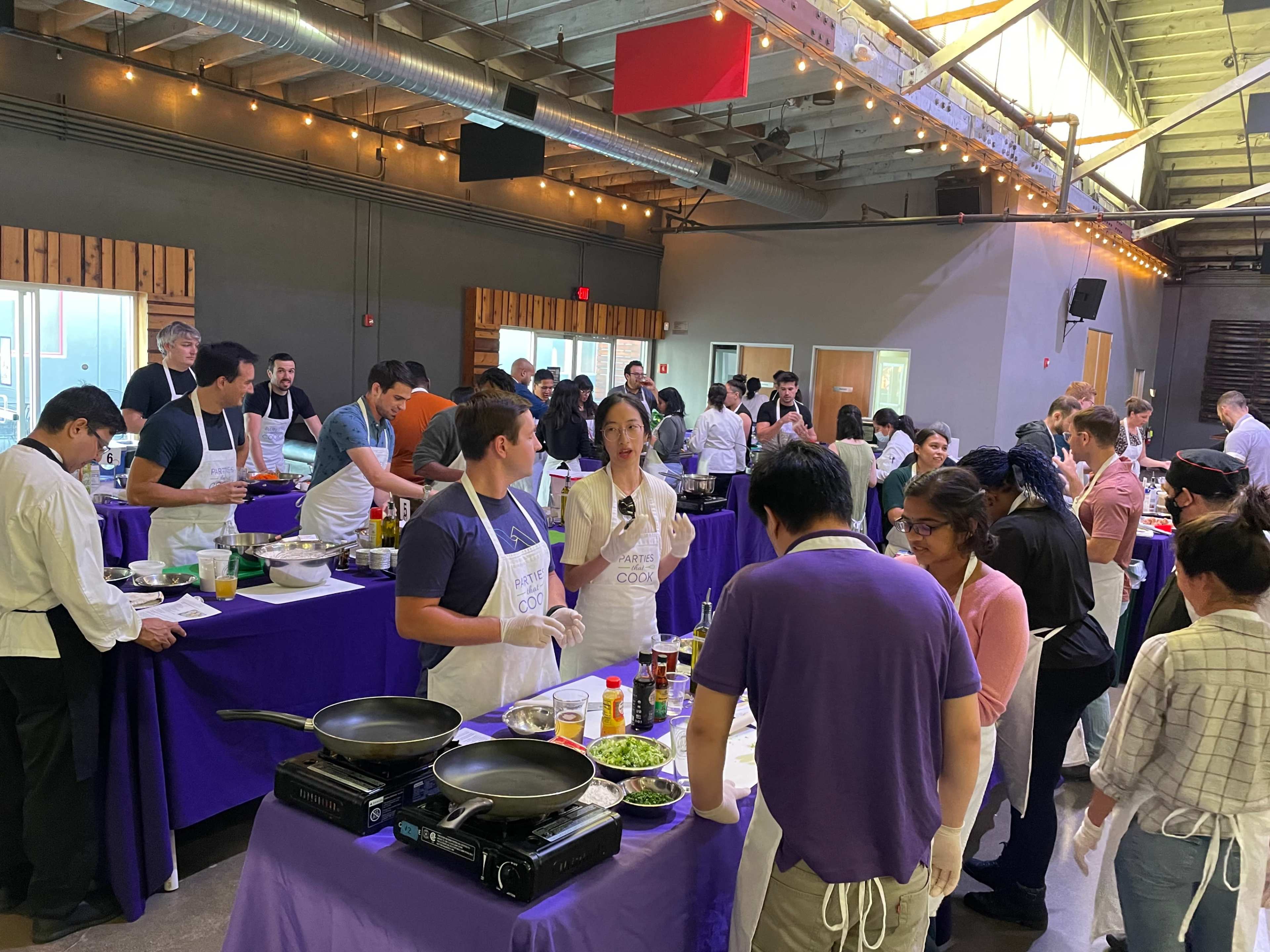 A large cooking event takes place in a spacious venue with participants gathered around tables, preparing various dishes.