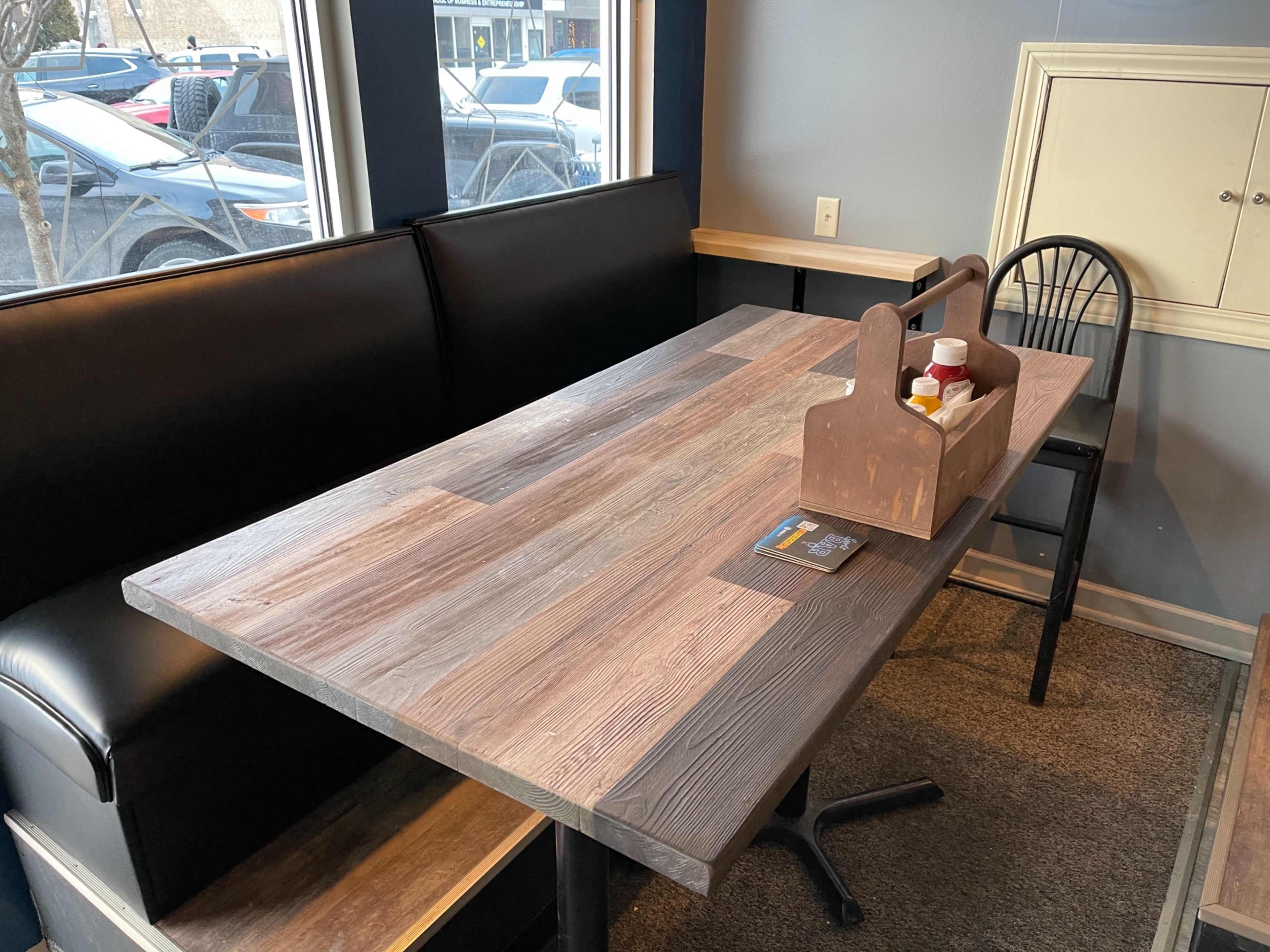 A wooden table with a carrying caddy, surrounded by a black bench and a chair, is positioned near a window in a restaurant.