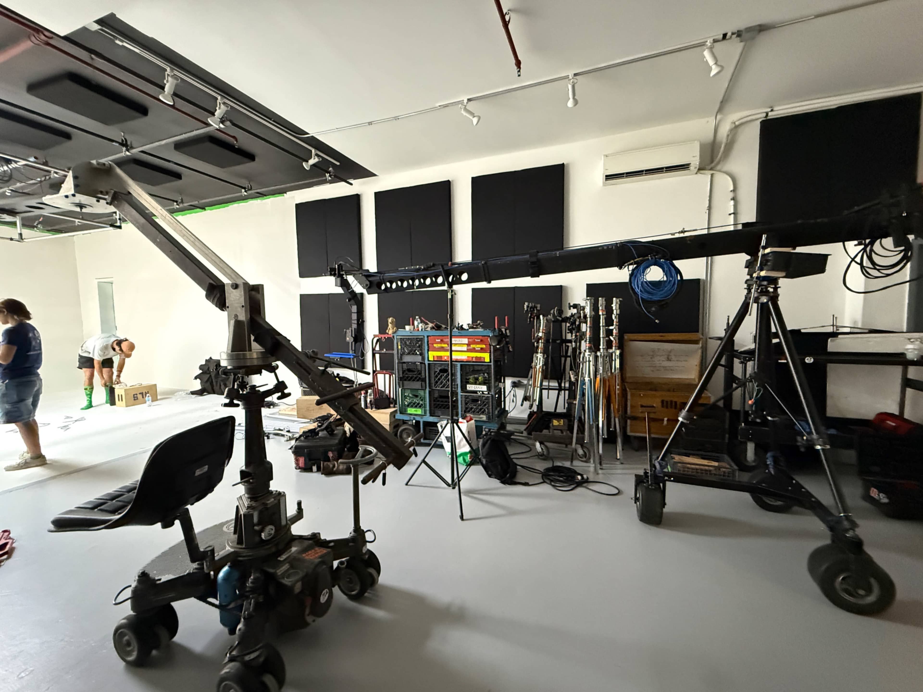 The image shows a film studio with various equipment, including a camera crane and lighting setup, along with crew members preparing the space.