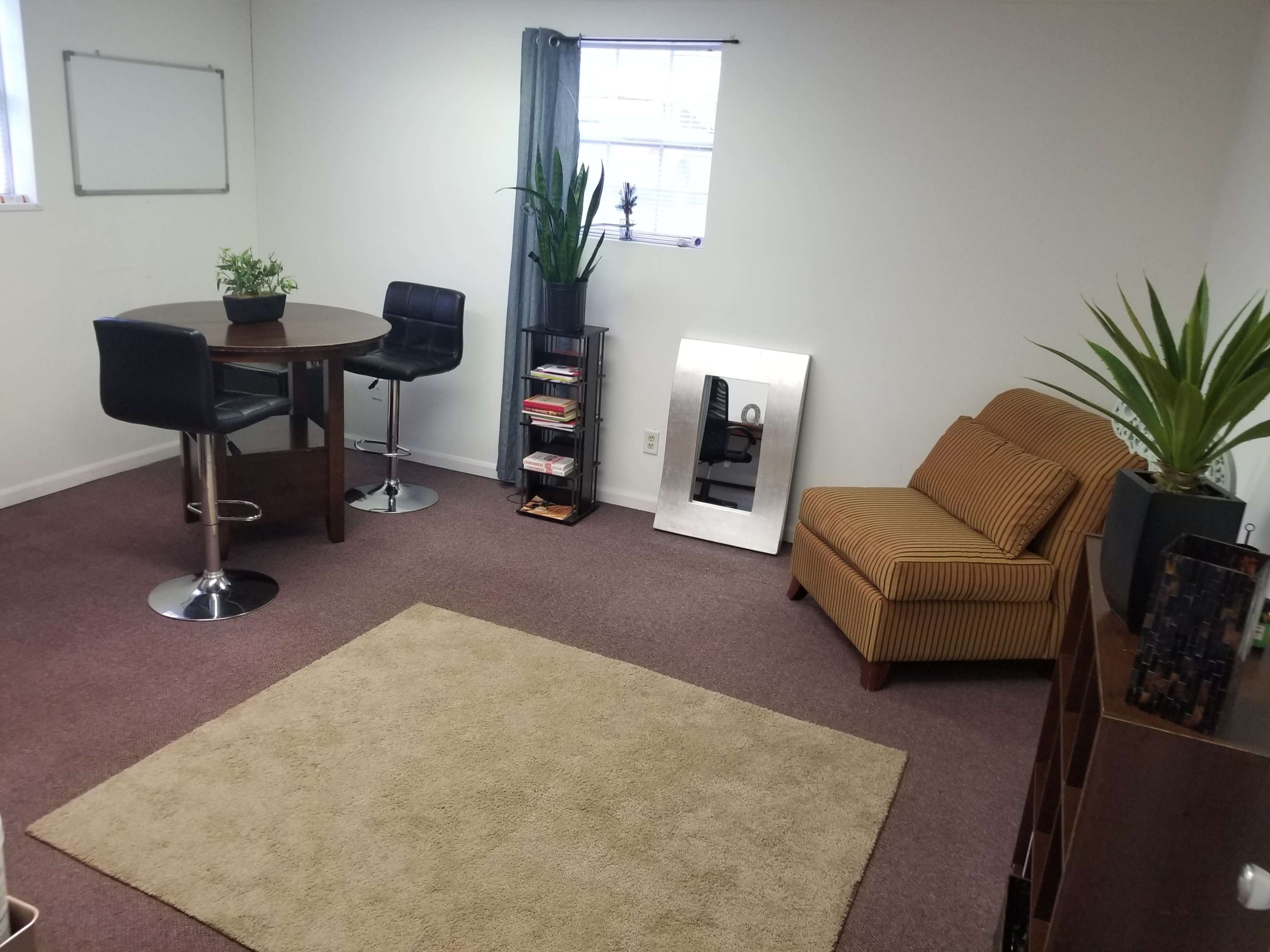 A sparsely furnished room with a round table, two black stools, a couch, a mirror, and a small bookshelf, all arranged around a beige rug.