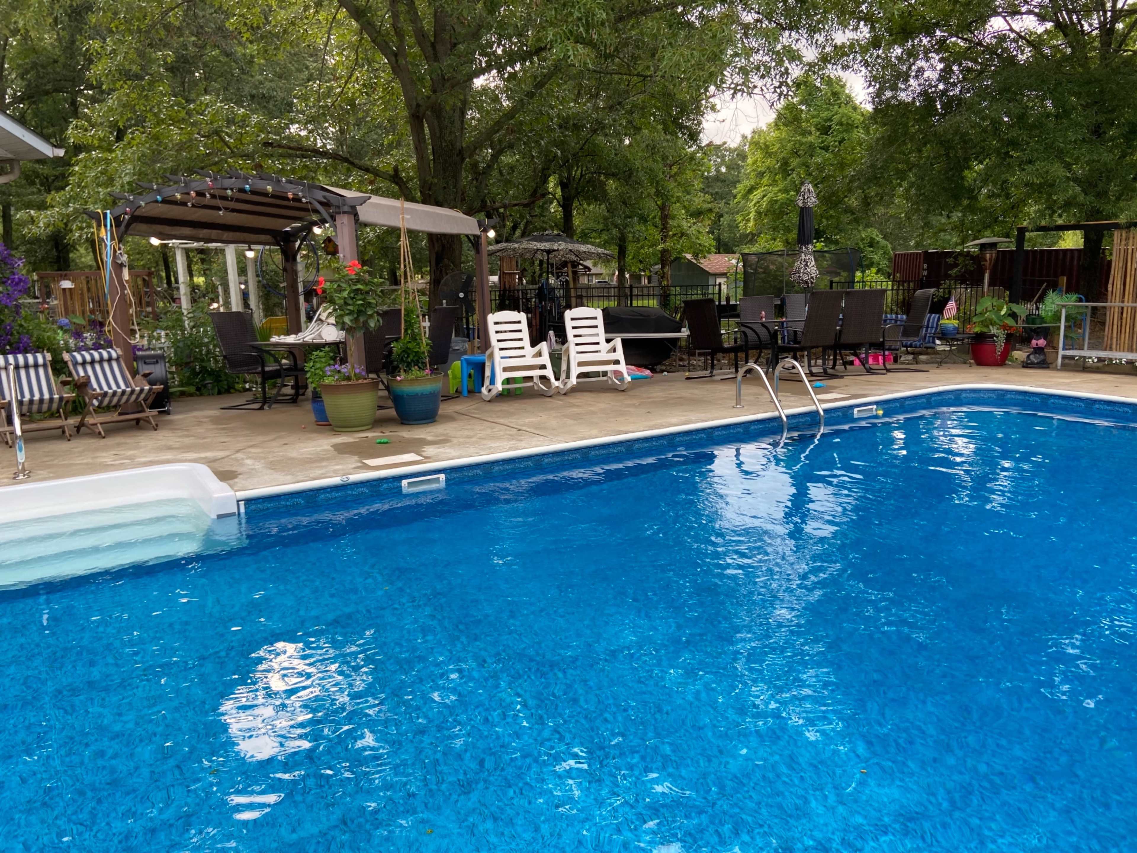A clear blue swimming pool surrounded by lounge chairs, potted plants, and a shaded seating area with a pergola.